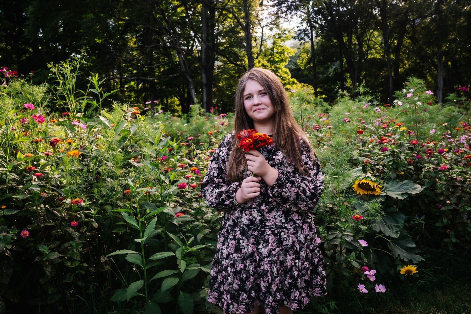 Young girl in floral dress holding red flowers in a lush garden with colorful blooms and trees.