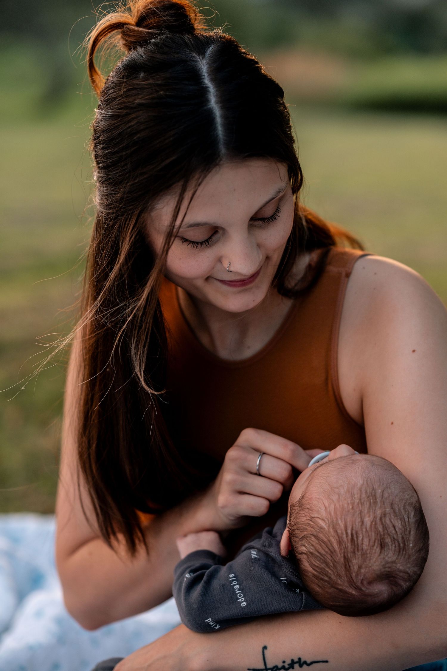 A dark-haired woman in an orange tank top tenderly cradles her sleeping newborn baby outdoors.