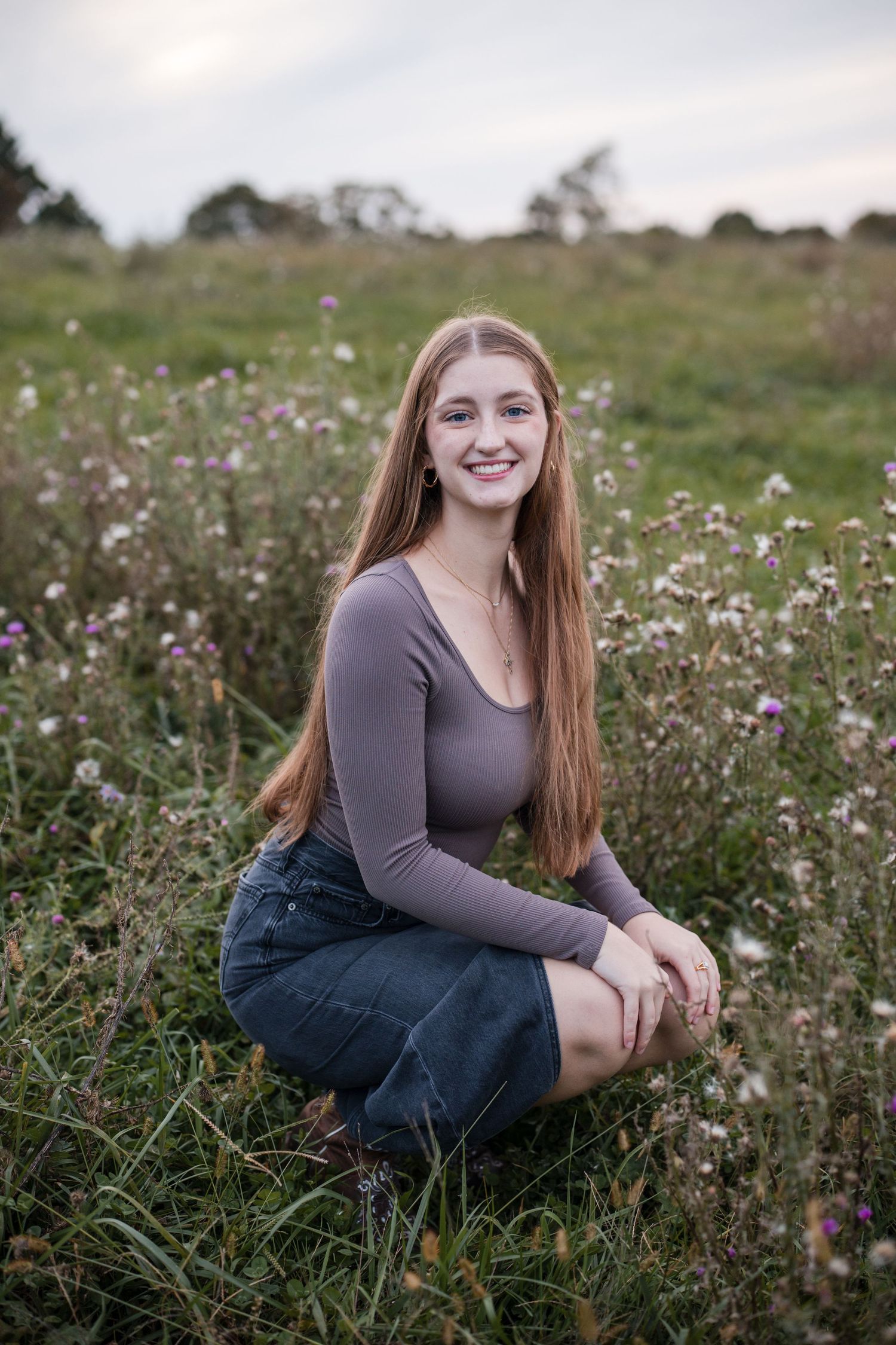 Young woman with long red hair smiling in a wildflower field, wearing a gray top and denim skirt.