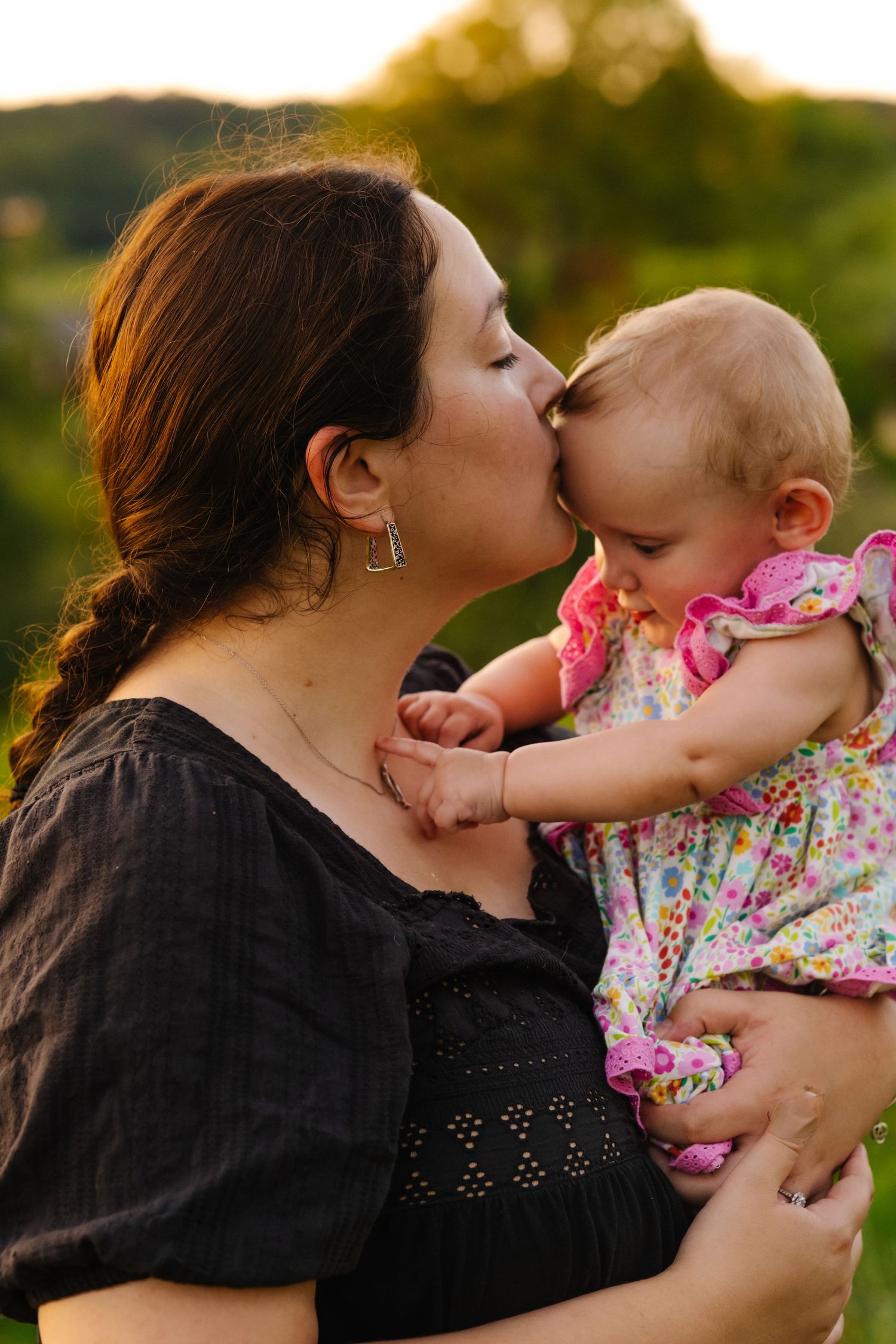 A mother tenderly kisses her baby girl outdoors at golden hour, both dressed in black and floral outfits.