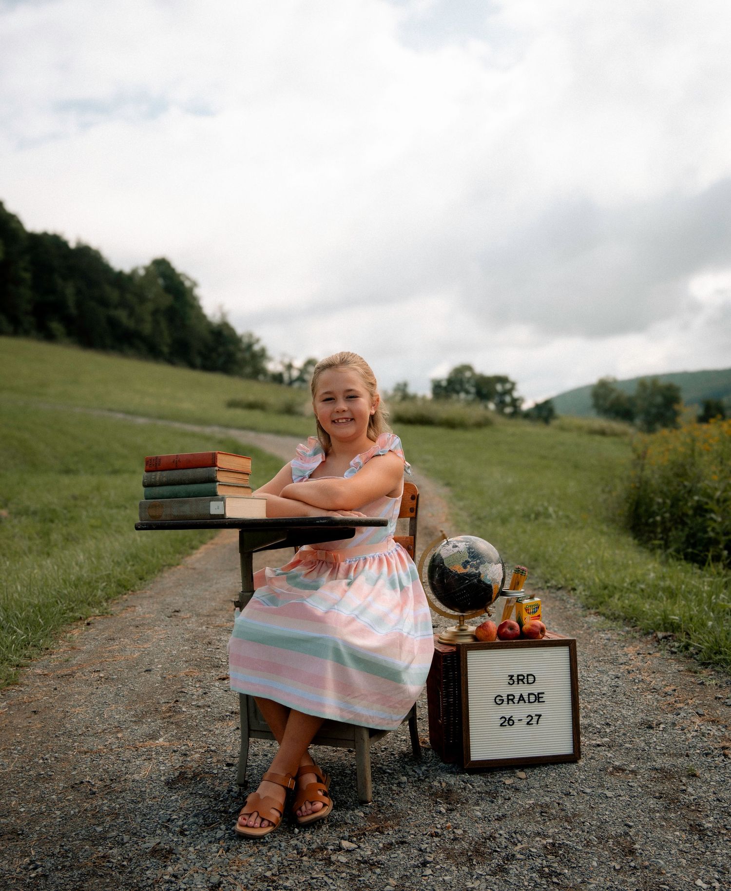 A smiling girl in a pink dress sits at a school desk outdoors holding a 3rd grade sign, books, and a globe.