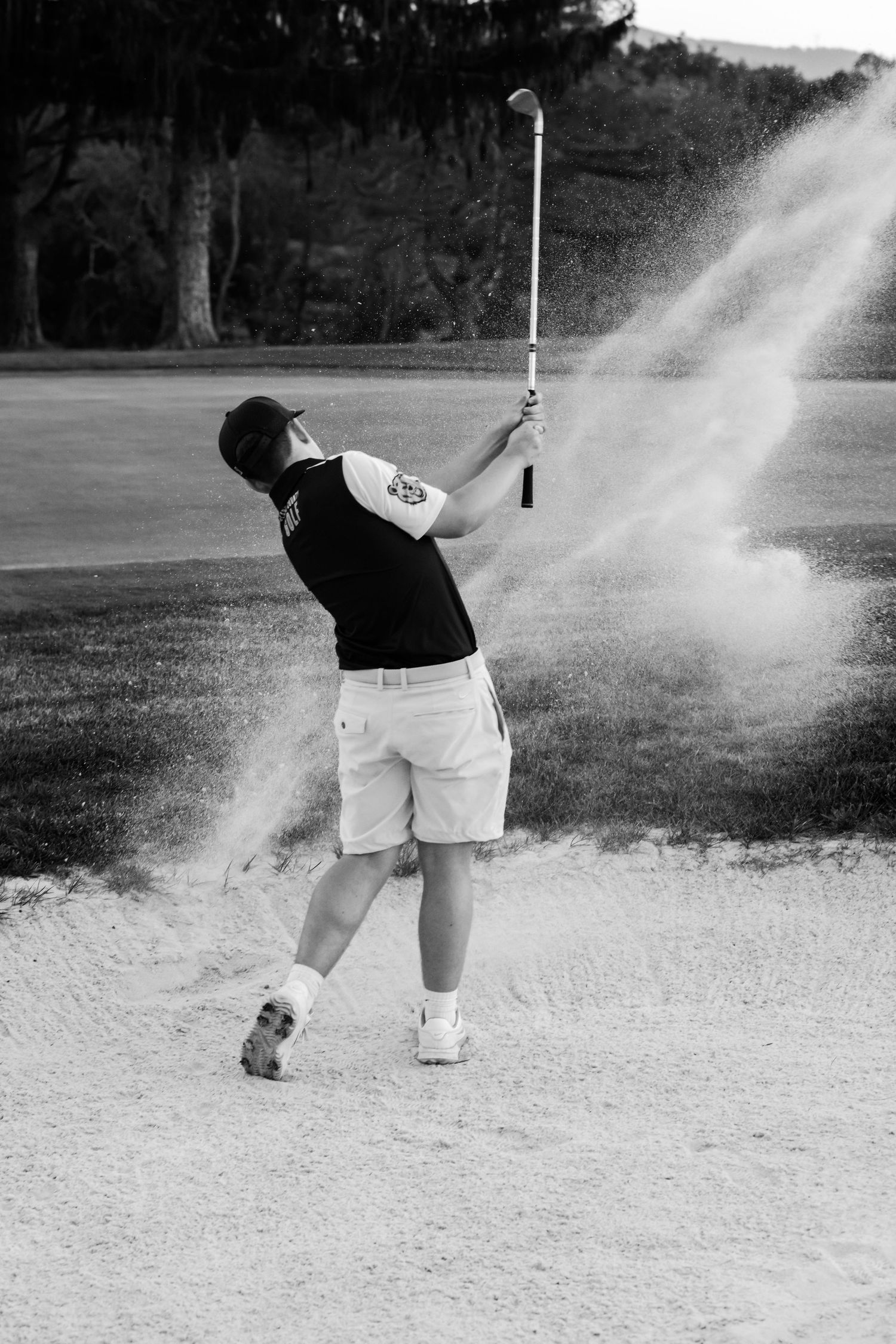 Black and white photo of a golfer executing a sand bunker shot, sending sand flying on a golf course.