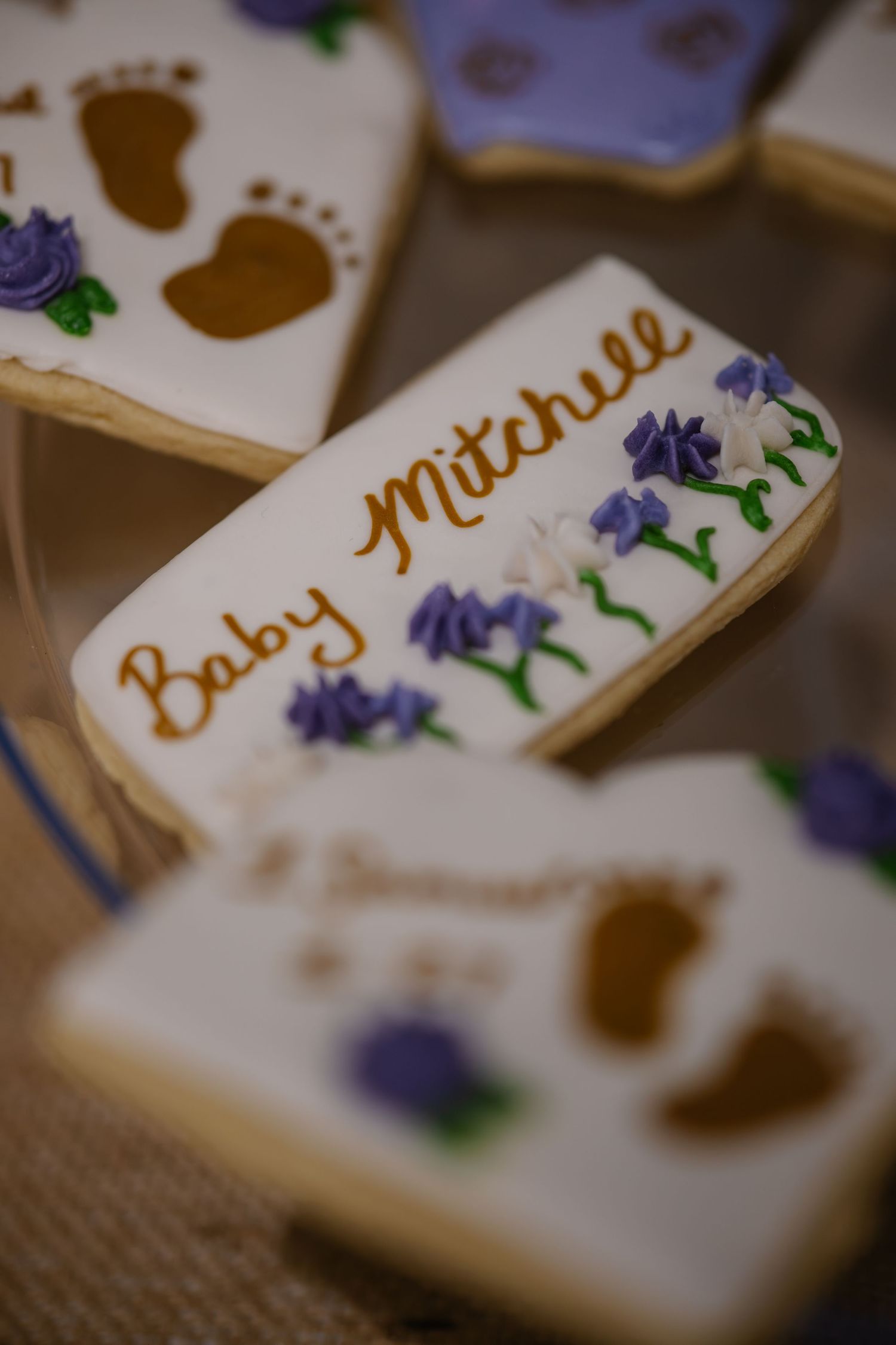 Decorated sugar cookies reading Baby Mitchell with purple floral icing and baby footprint designs for a baby shower.