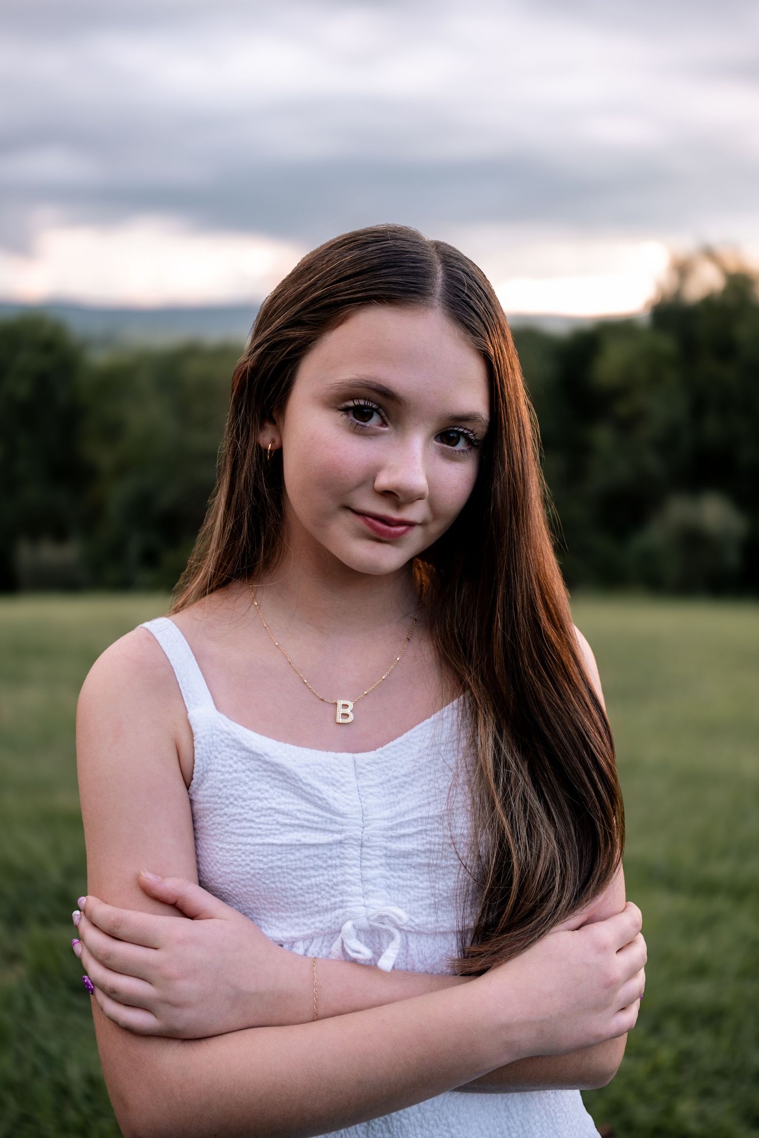 Teen girl with long brown hair in white dress holds arms crossed in grassy field.