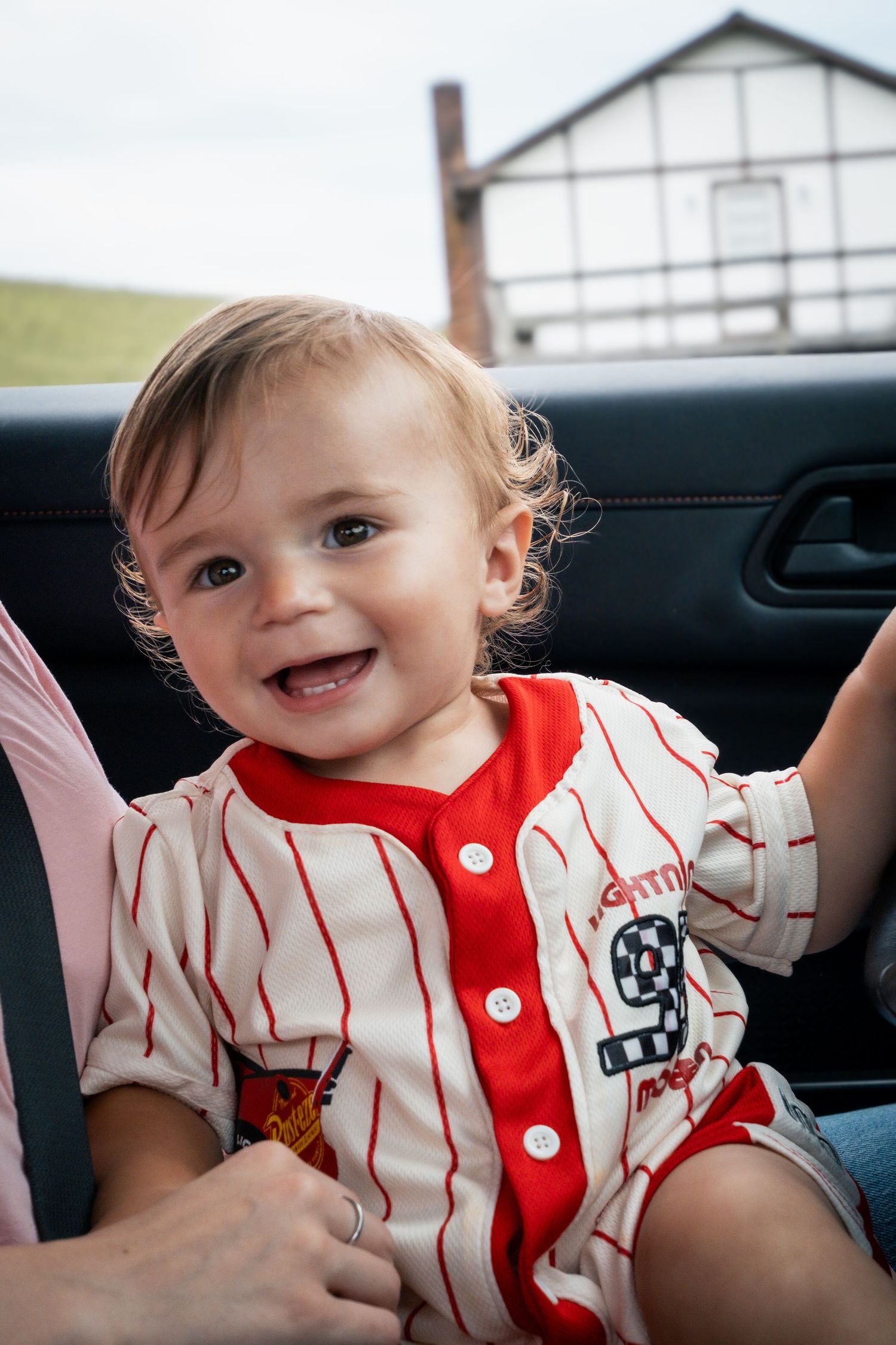 Smiling baby in red and white striped baseball outfit sitting in car seat with building visible through window.