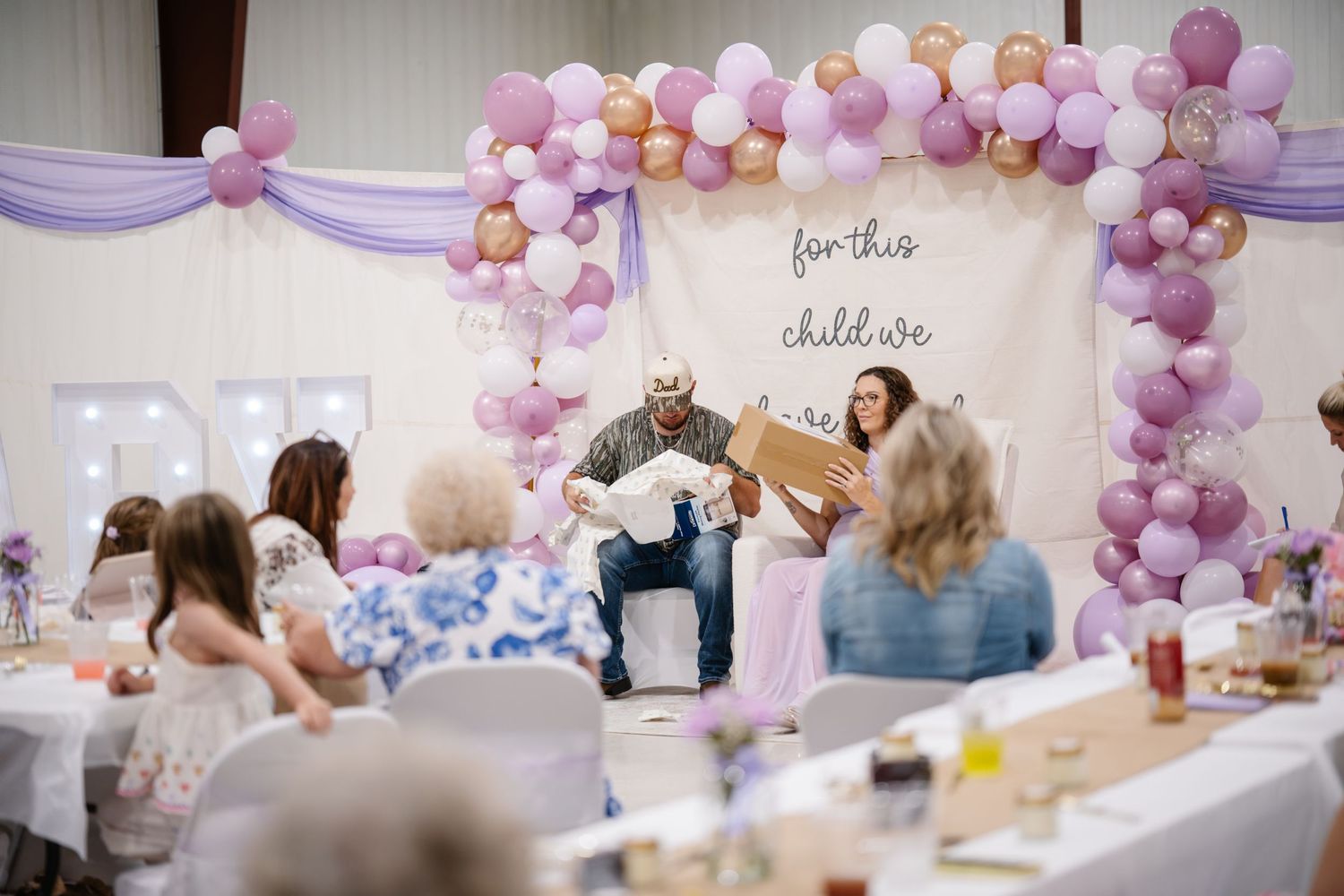 Baby shower celebration with purple balloon arch, guests seated at tables, and parents opening gifts in decorated venue.