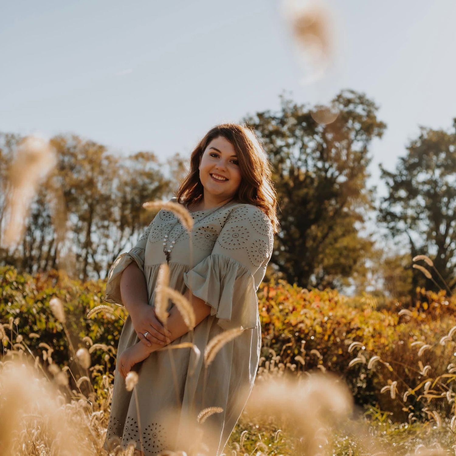 A smiling woman in a sage green dress poses in a sunlit field with tall grasses and autumn trees in the background.