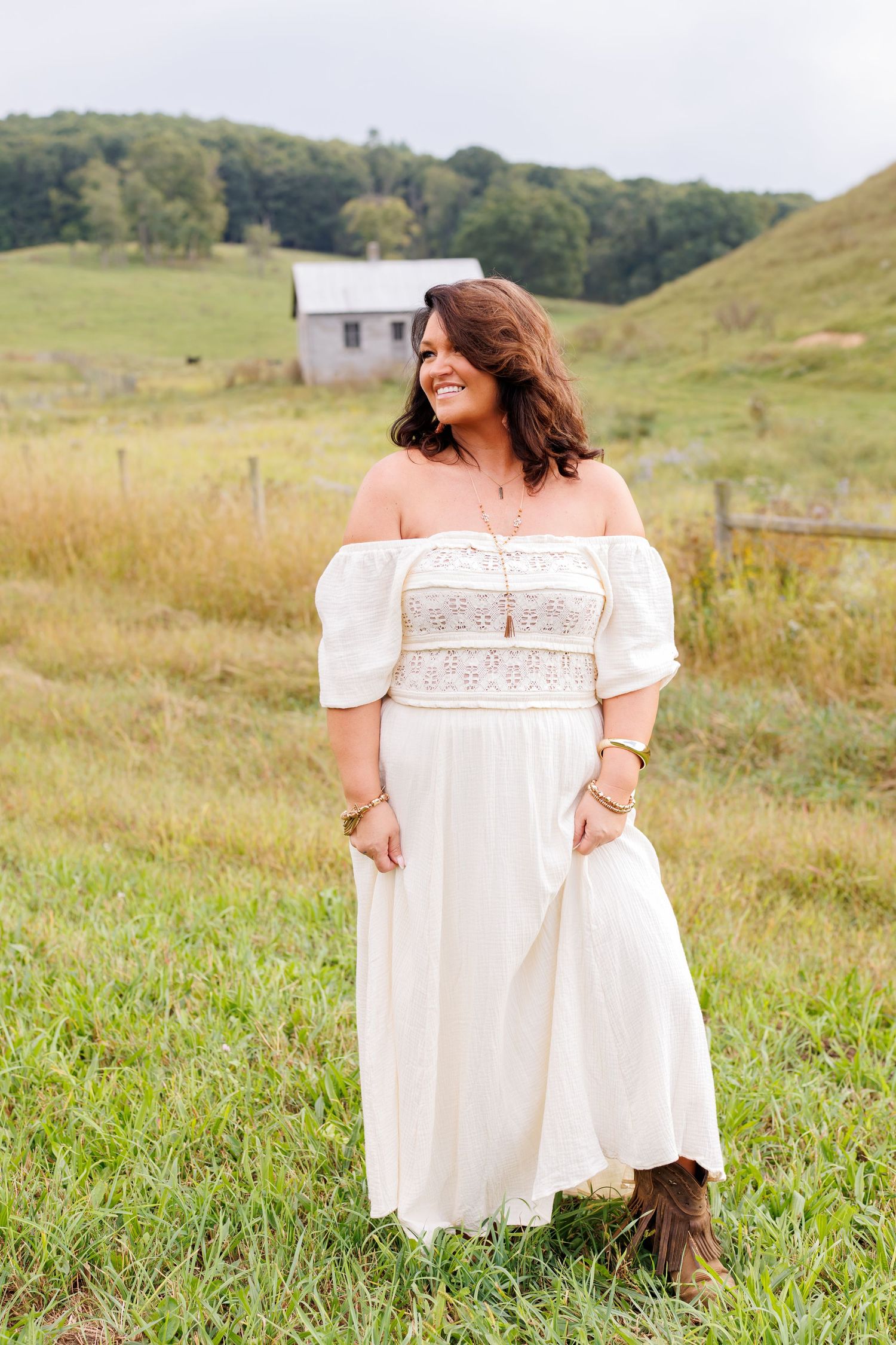 Woman in white off-shoulder boho dress and cowboy boots smiling in a green field with a rustic barn backdrop.