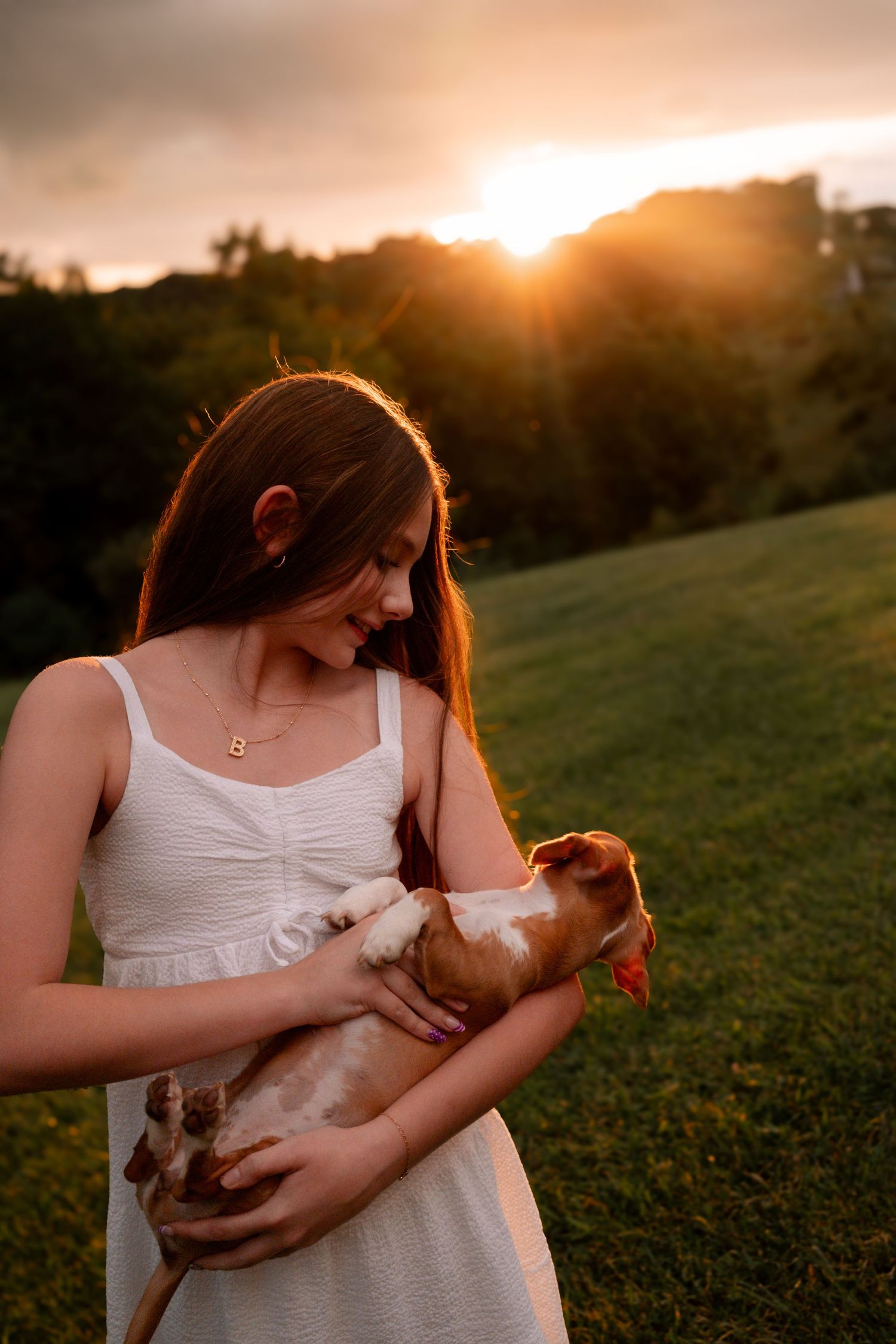 Smiling teenage girl in white dress holds small brown dog during golden hour sunset outdoors.