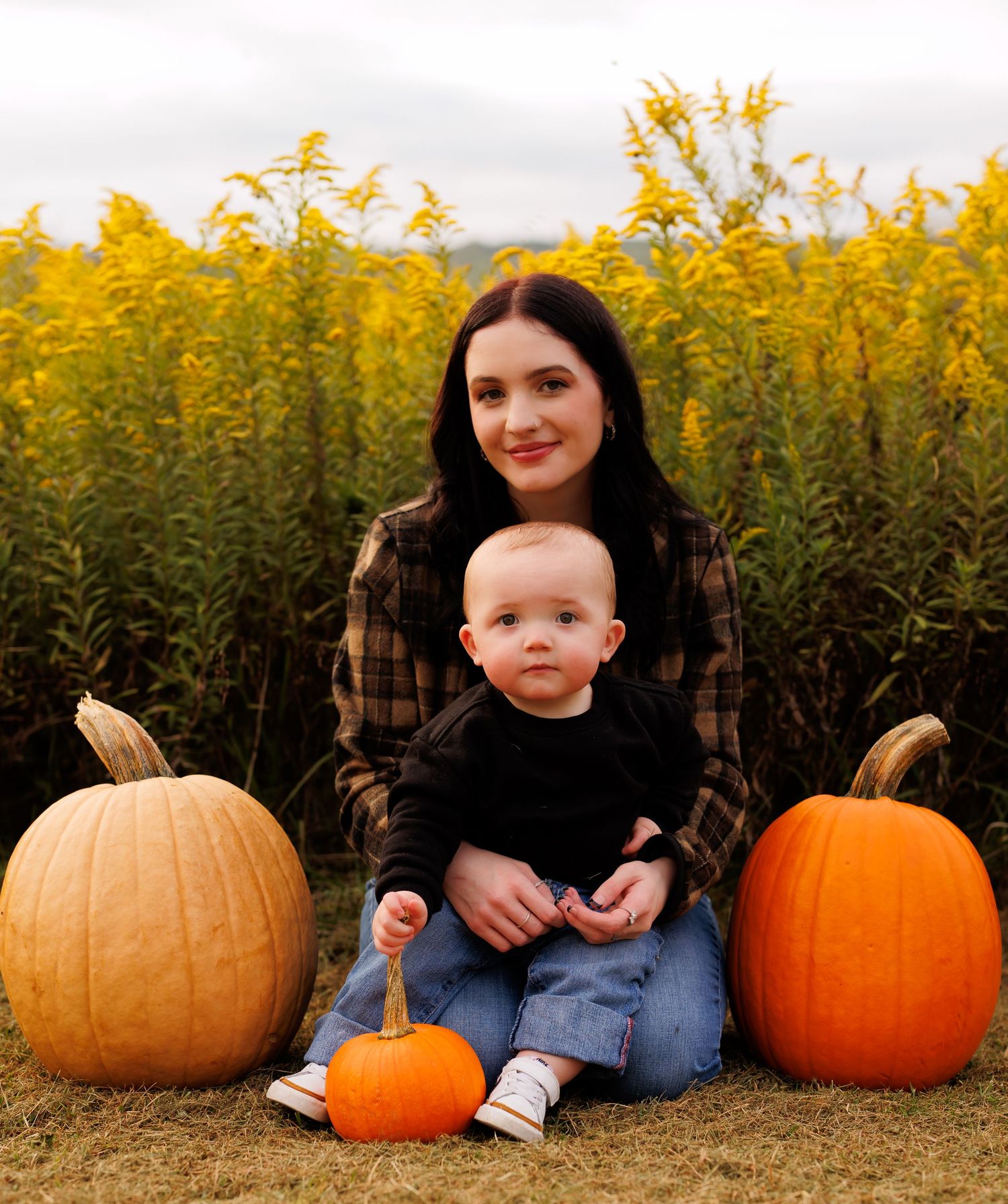 A young woman and baby sit among orange pumpkins at a fall pumpkin patch with golden wildflowers behind them.