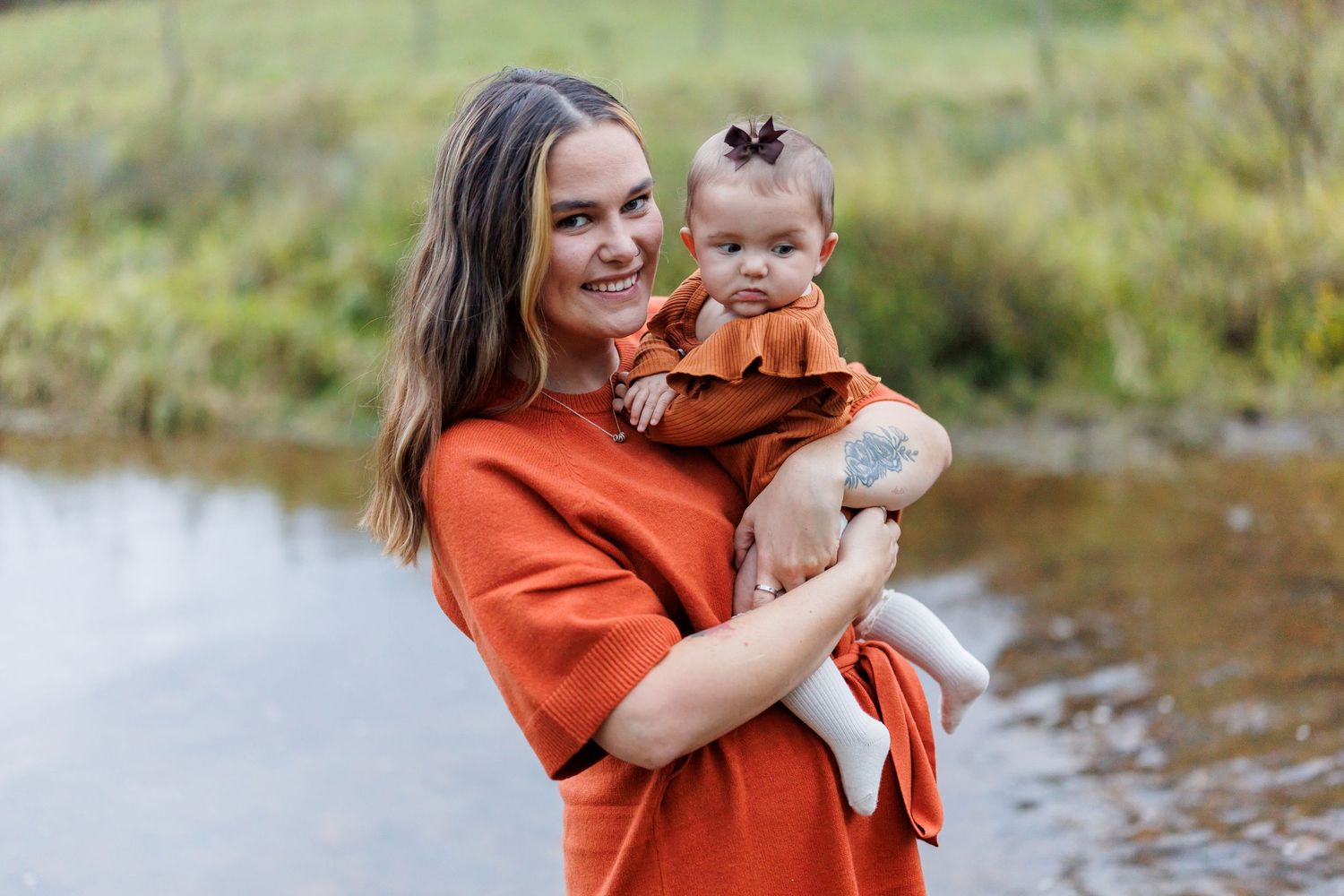 A smiling young mother in an orange dress holds her baby girl near a serene creek outdoors.