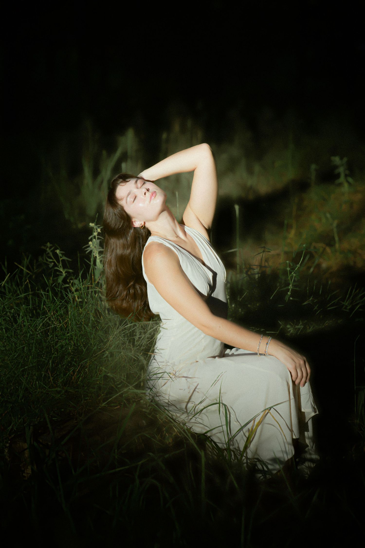 A woman in a white dress sits in tall dark grass at night illuminated by dramatic lighting creating an ethereal mood.