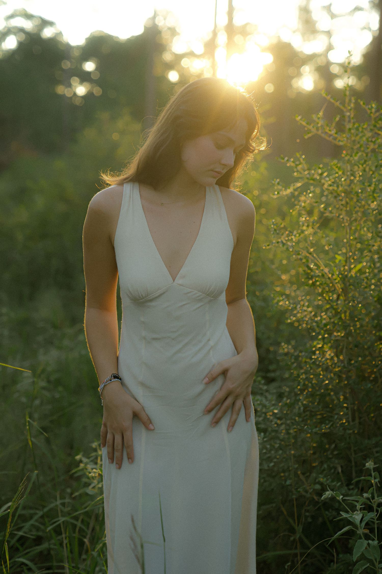 A woman in a white dress stands in a field at sunset with golden sunlight filtering through trees in the background.