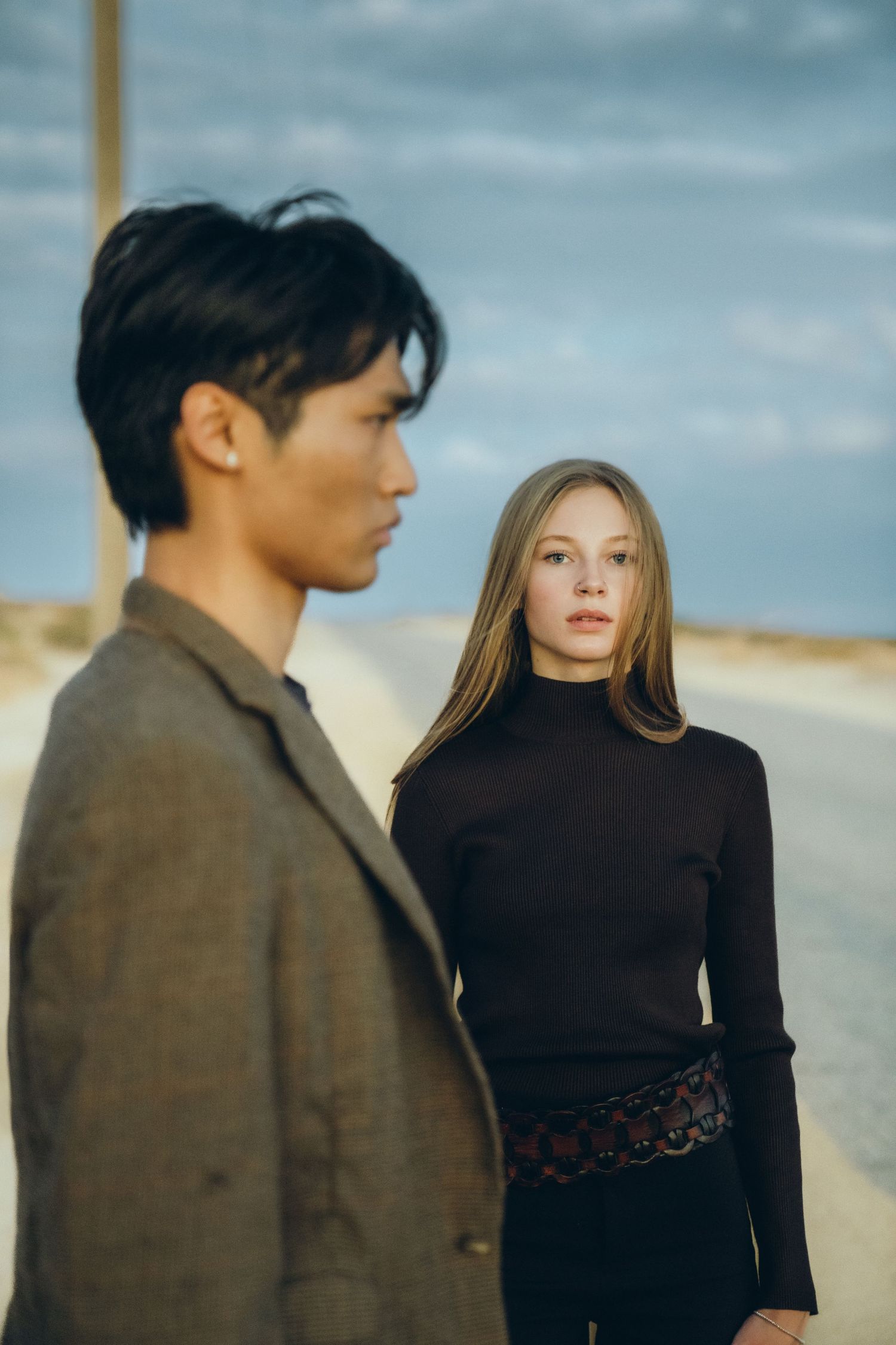 A couple in black and brown outfits stands against a cloudy sky backdrop by the seaside.