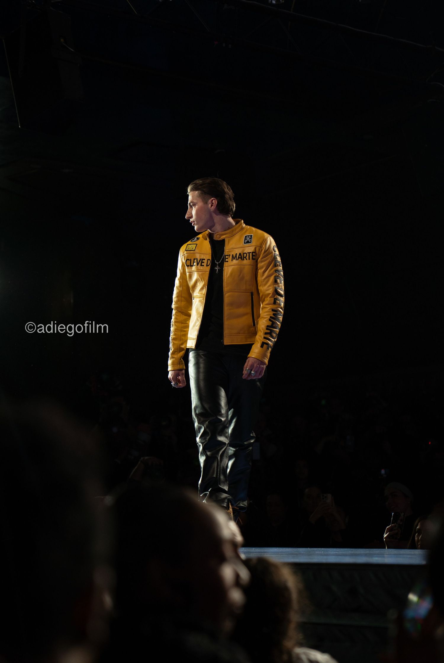 Performer in yellow jacket on dark stage with dramatic lighting and audience silhouettes.