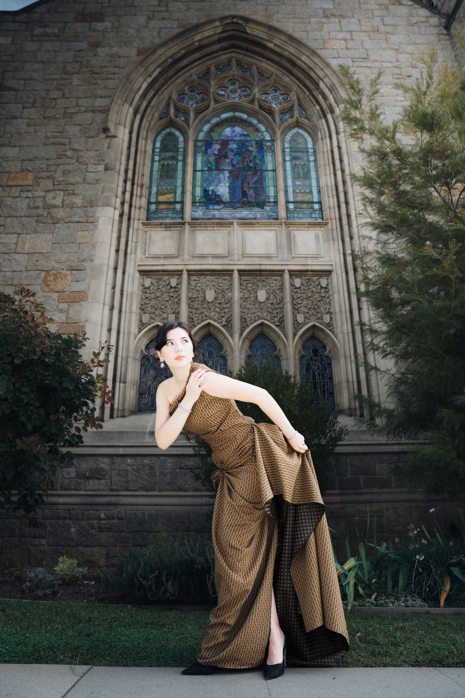 A figure in a brown vintage dress poses dramatically against a stone church wall with stained glass windows.