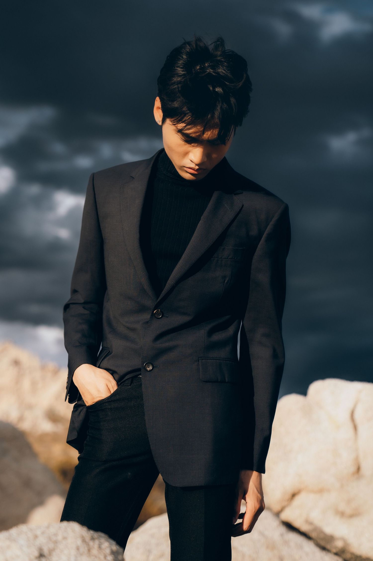 Young man in all-black suit and turtleneck poses outdoors against dramatic cloudy sky and rocky landscape.