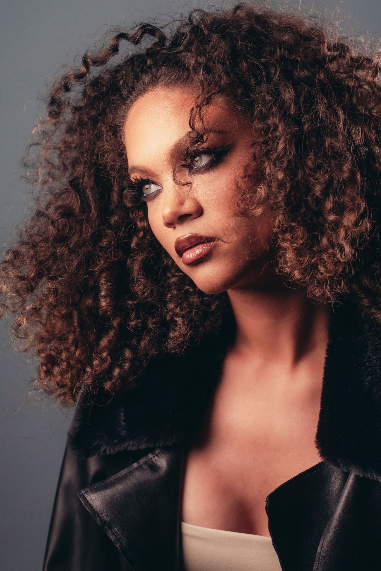 A woman with voluminous curly hair wearing a leather jacket, showcasing bold eye makeup against a gray background.