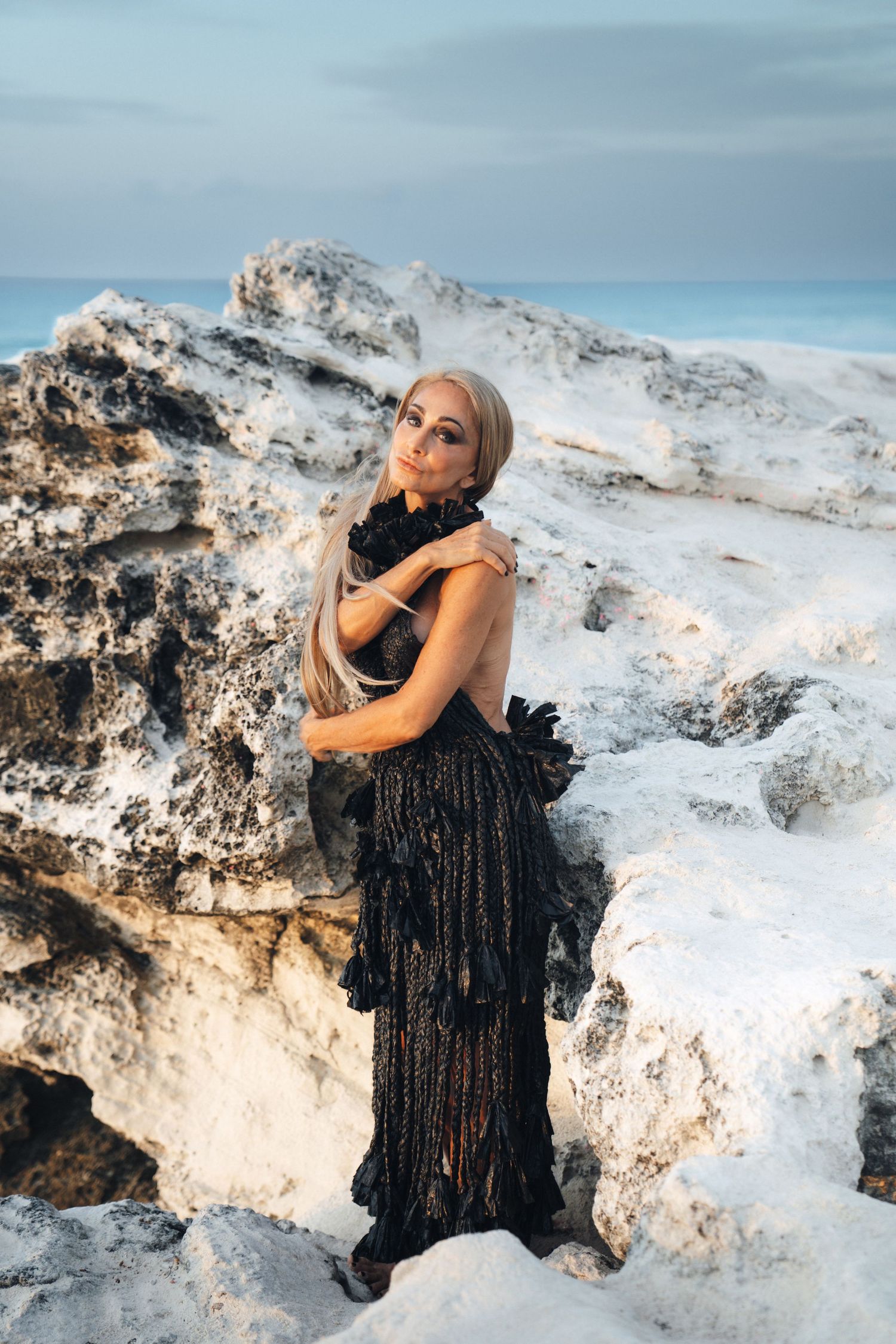 A person in an elegant black gown poses dramatically among snowy white rock formations at sunset.