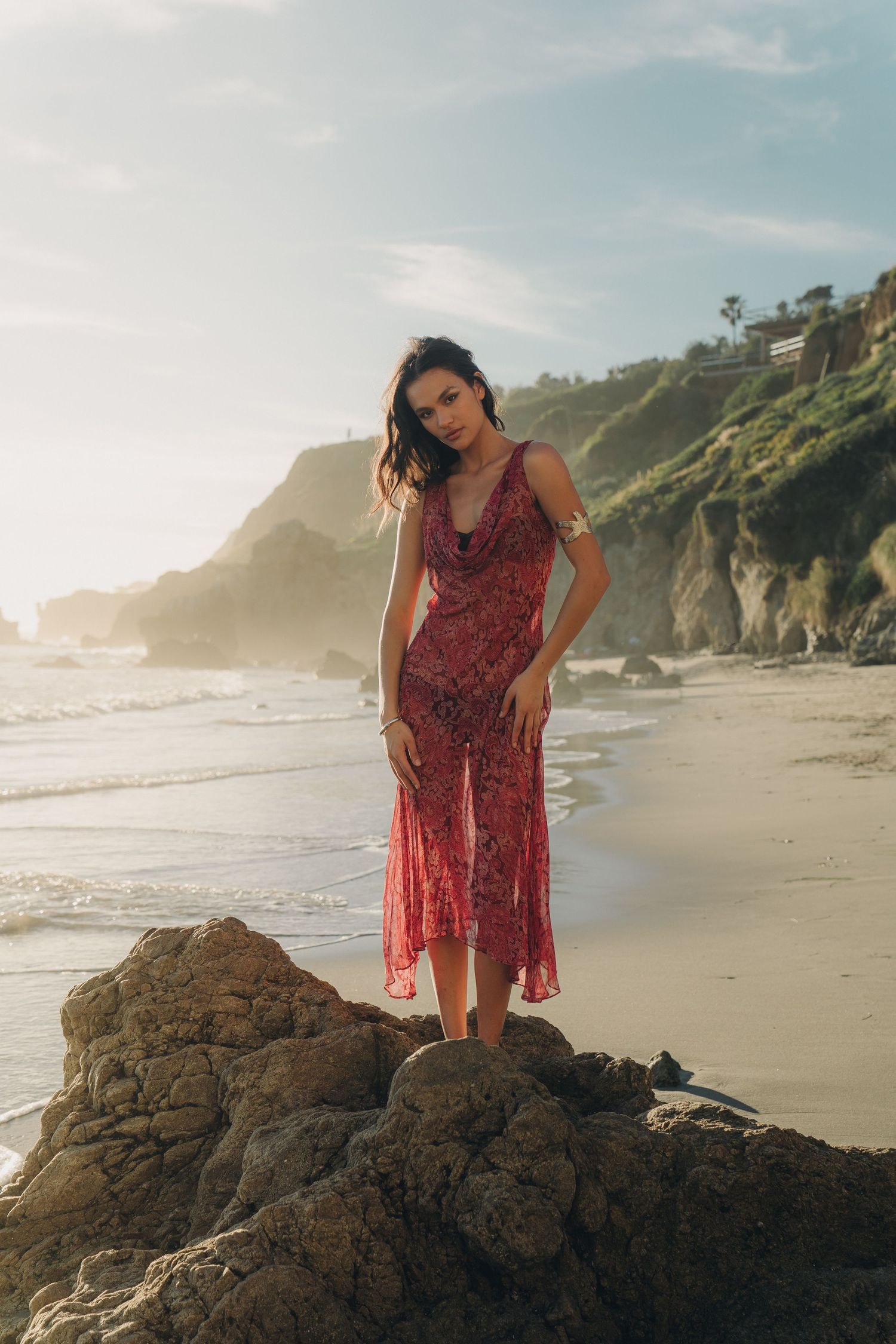 A person in a burgundy dress stands on coastal rocks at sunset while waves crash on a scenic beach.