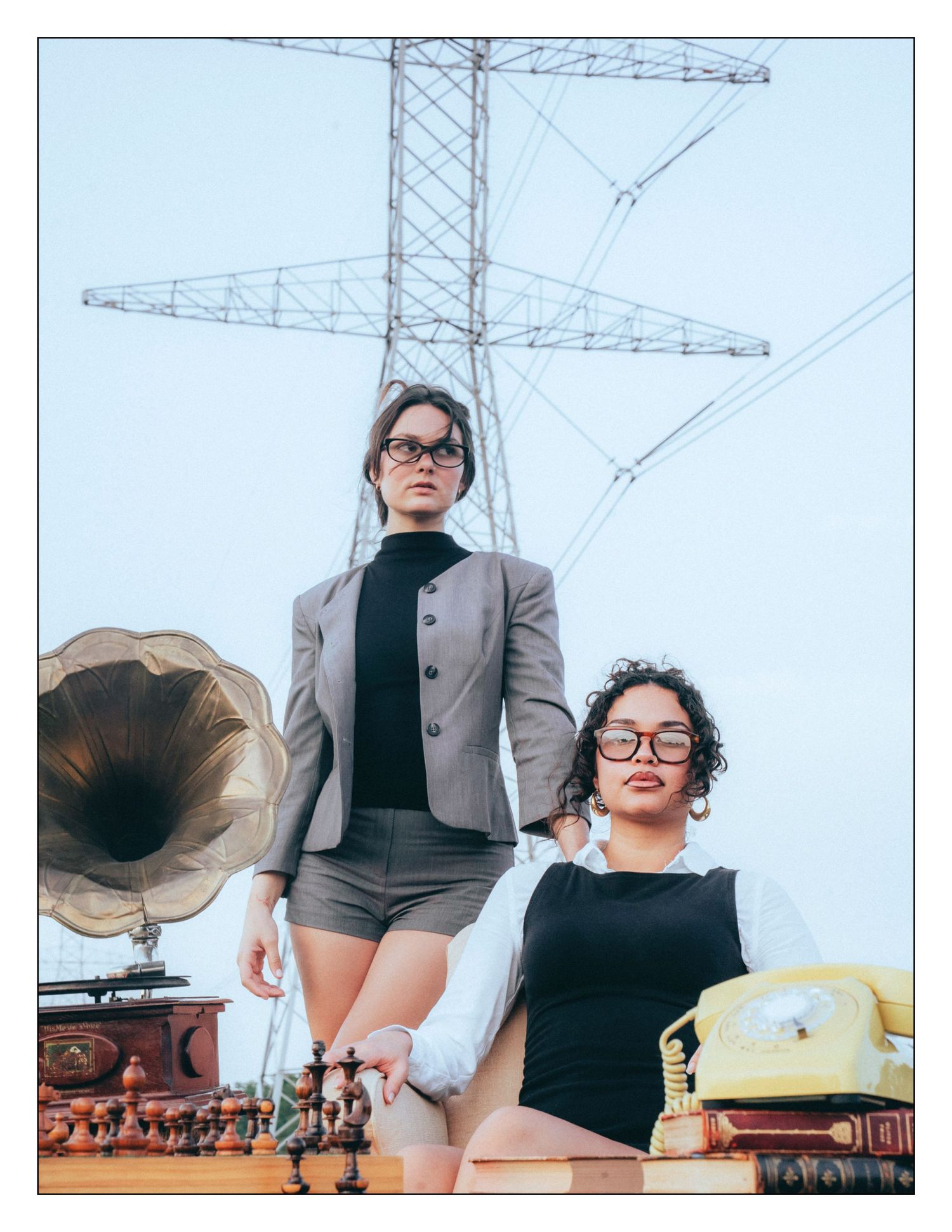 Two people stand in front of a vintage phonograph and rotary phone with an electrical tower in the background.