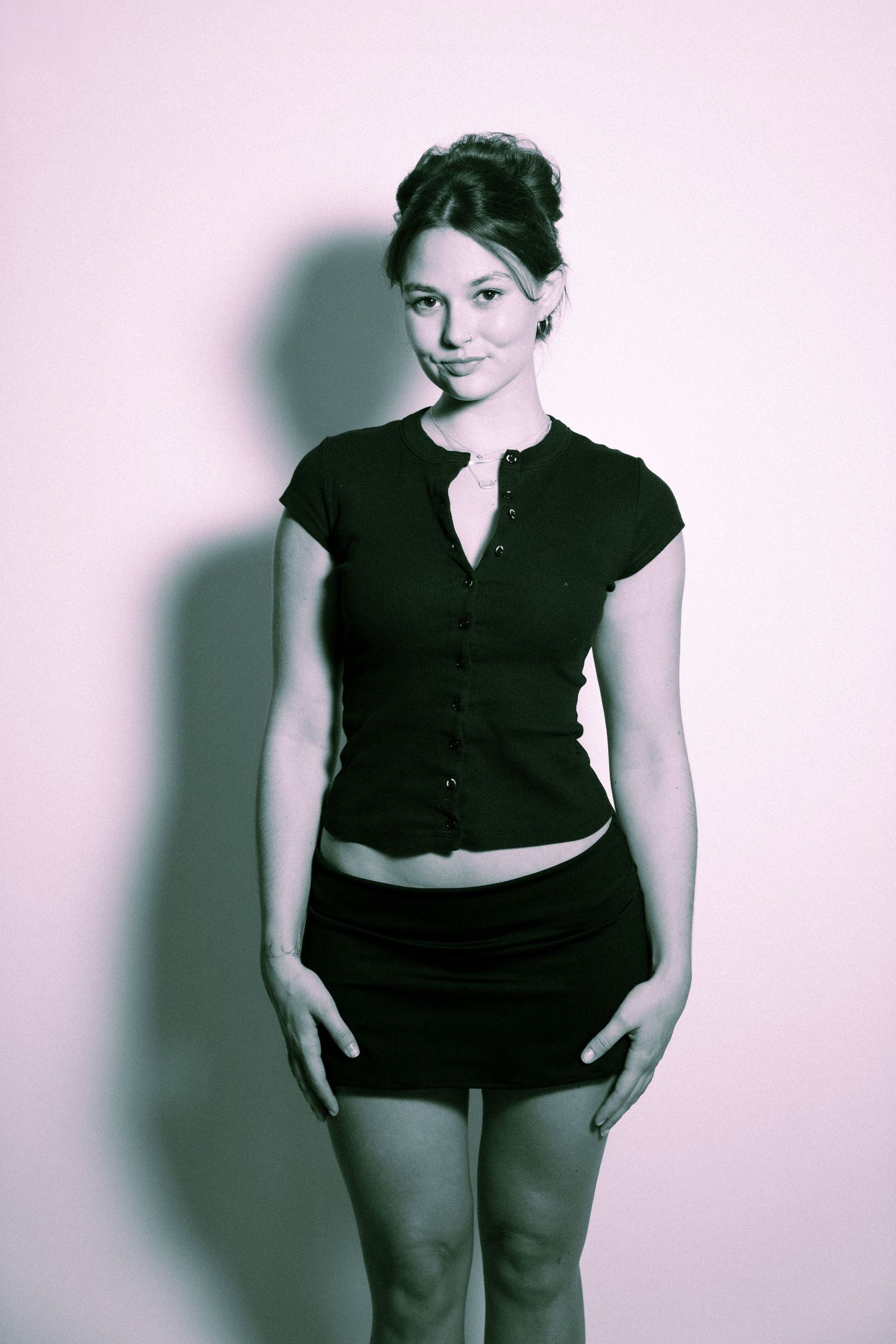 A person wearing a black short-sleeved button-up top and dark skirt poses against a pink backdrop.