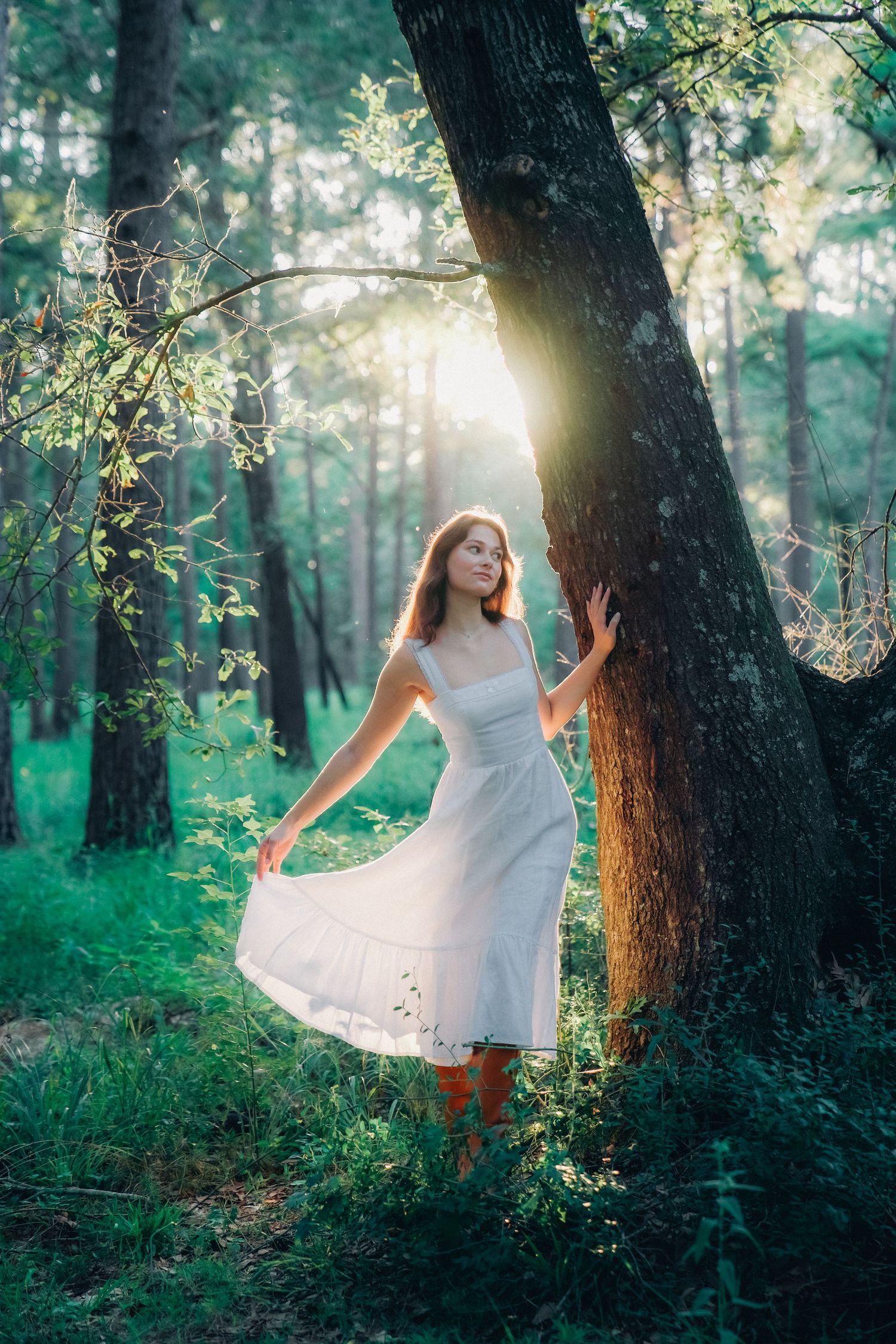 Whimsical forest portrait with sun flare and flowing white dress among green trees.
