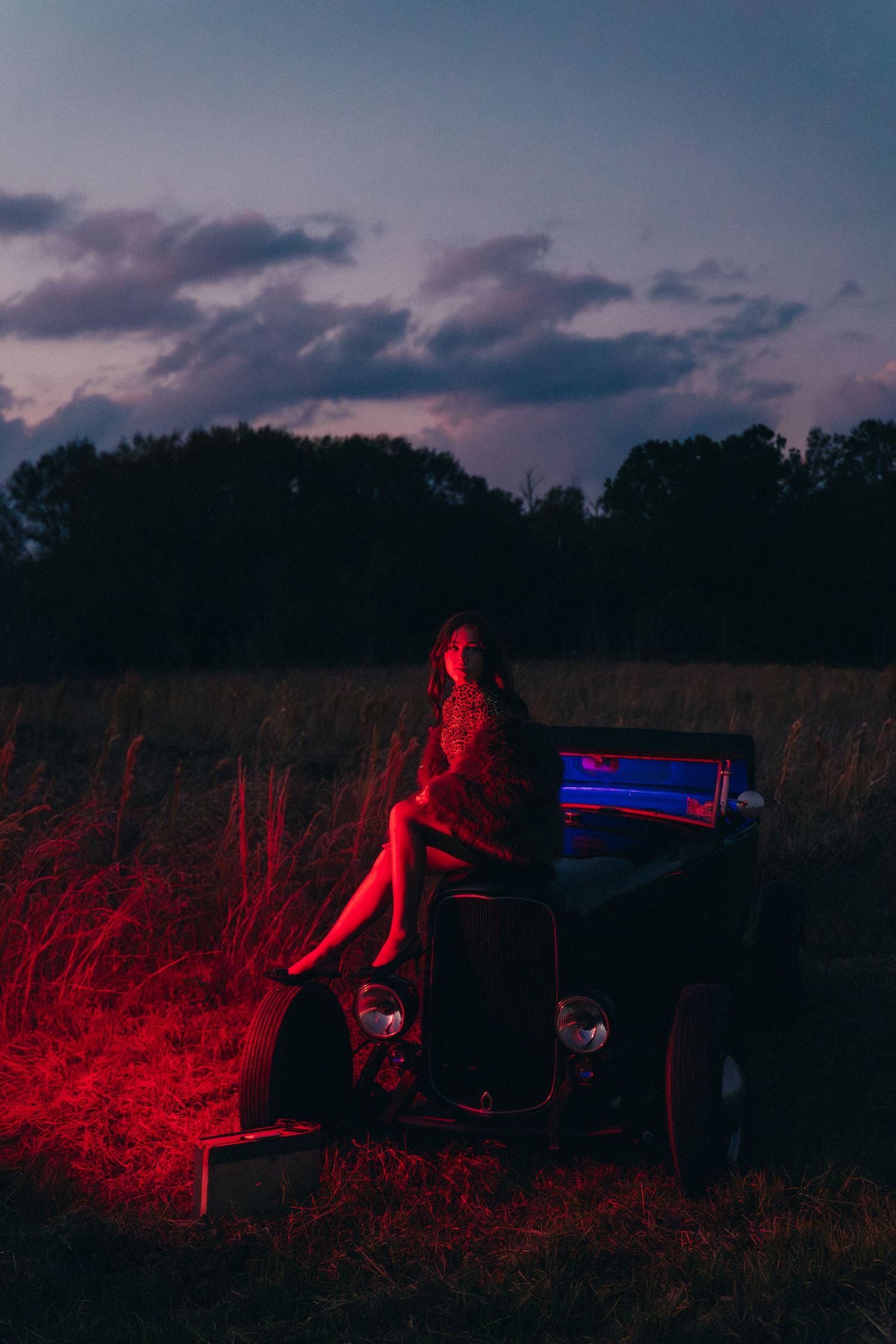 A figure in red lighting crouches in a dark field at dusk with dramatic moody sky and silhouetted trees in background.