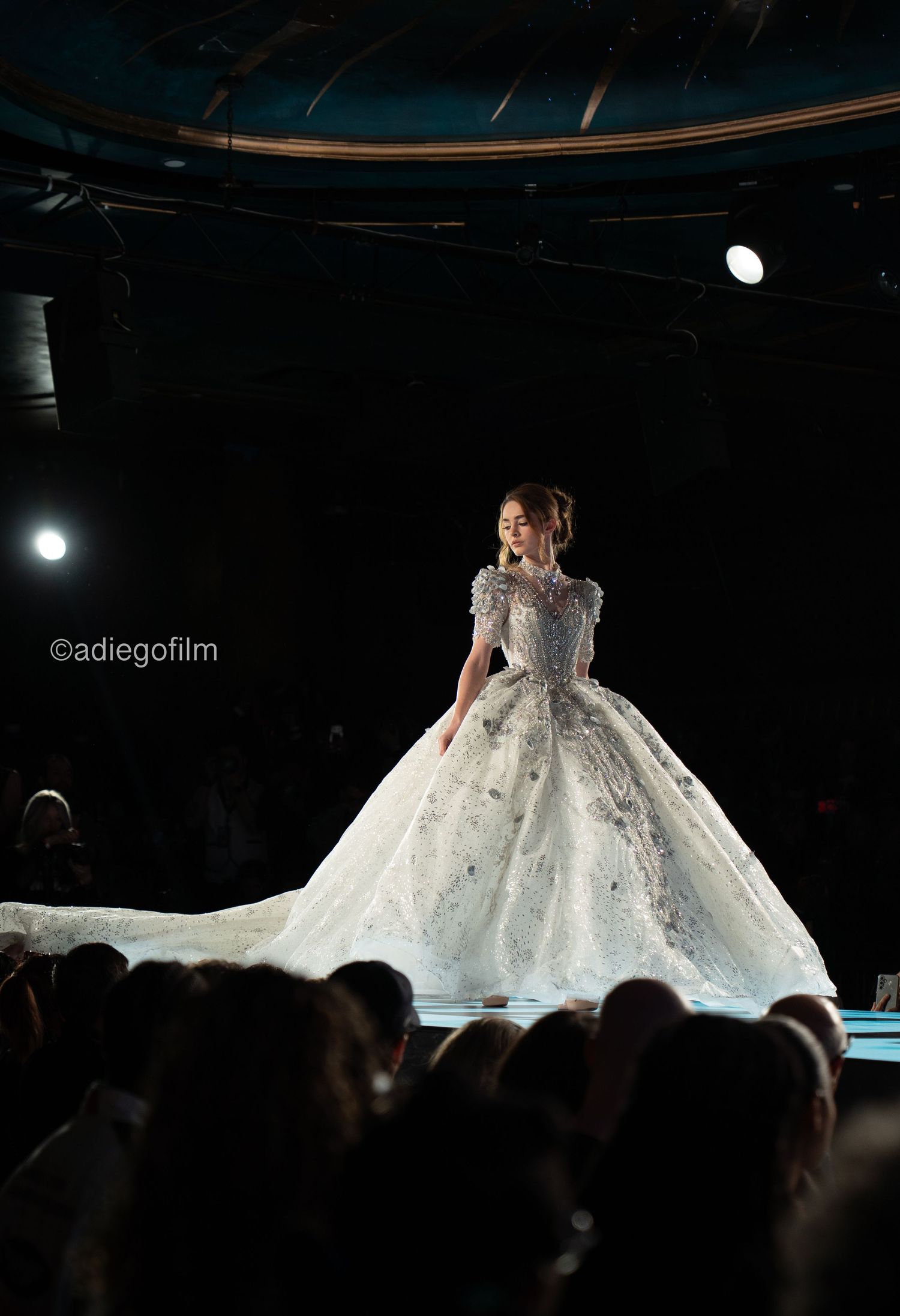 A model walks the runway in an elegant white ballgown with a dramatic full skirt during a fashion show.