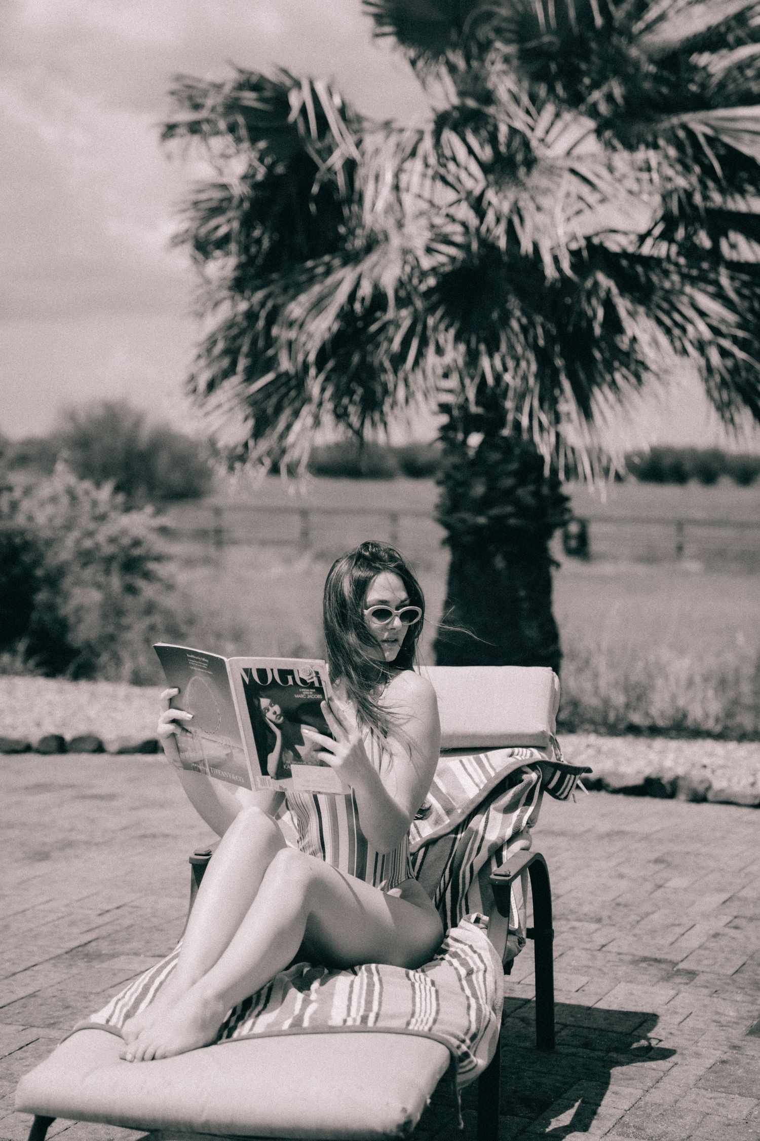 Woman relaxing on lounge chair reading magazine under palm trees in vintage black and white photograph.
