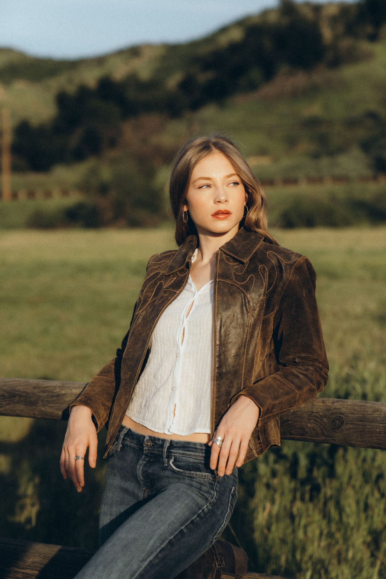 Young woman in brown suede jacket and jeans leaning on wooden fence with green hills in background.