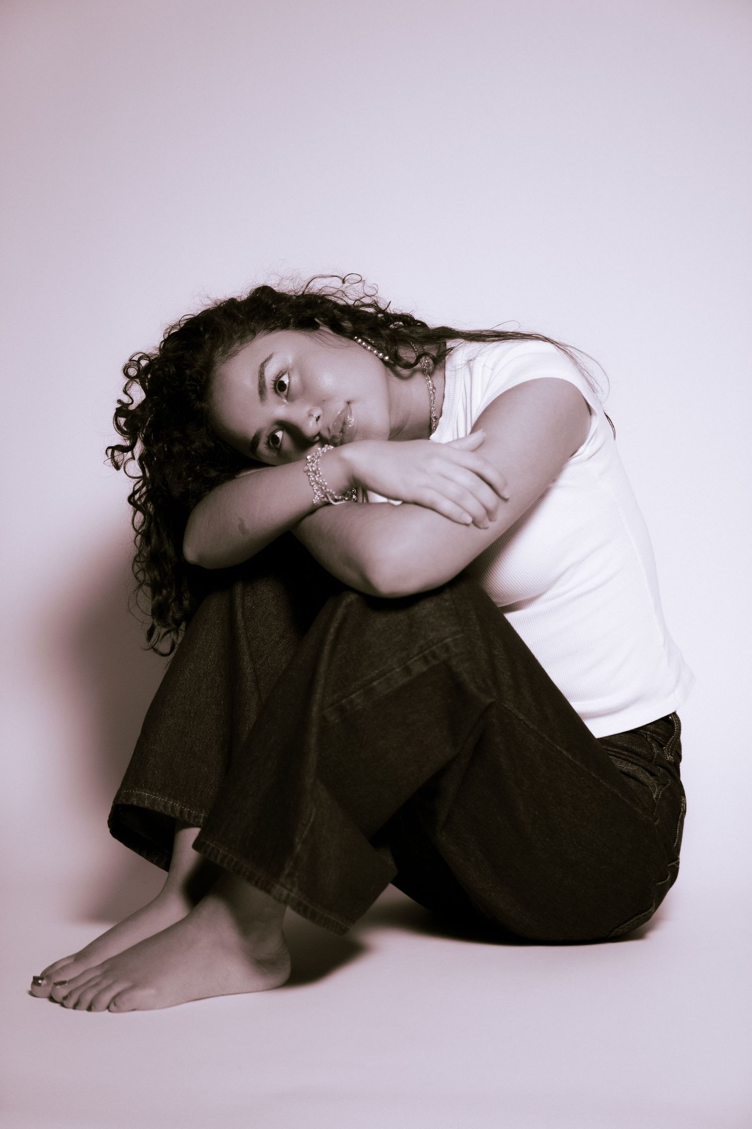 Portrait showing a seated figure in a white top and dark pants with curly hair, posed in a minimalist studio setting.