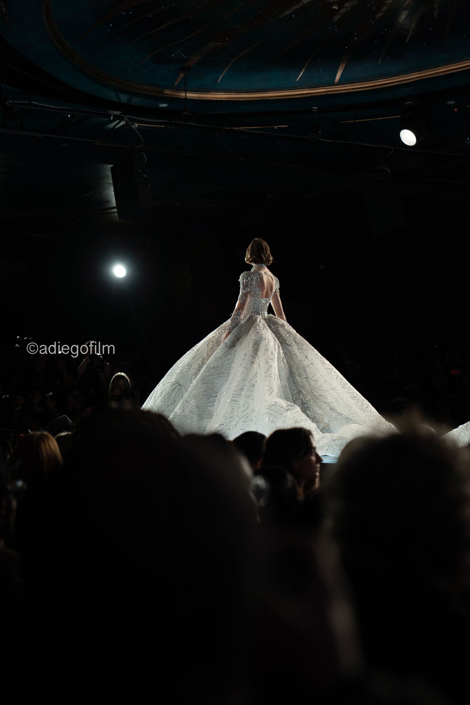 A model showcases a stunning white ballgown wedding dress on a dark runway with dramatic lighting.
