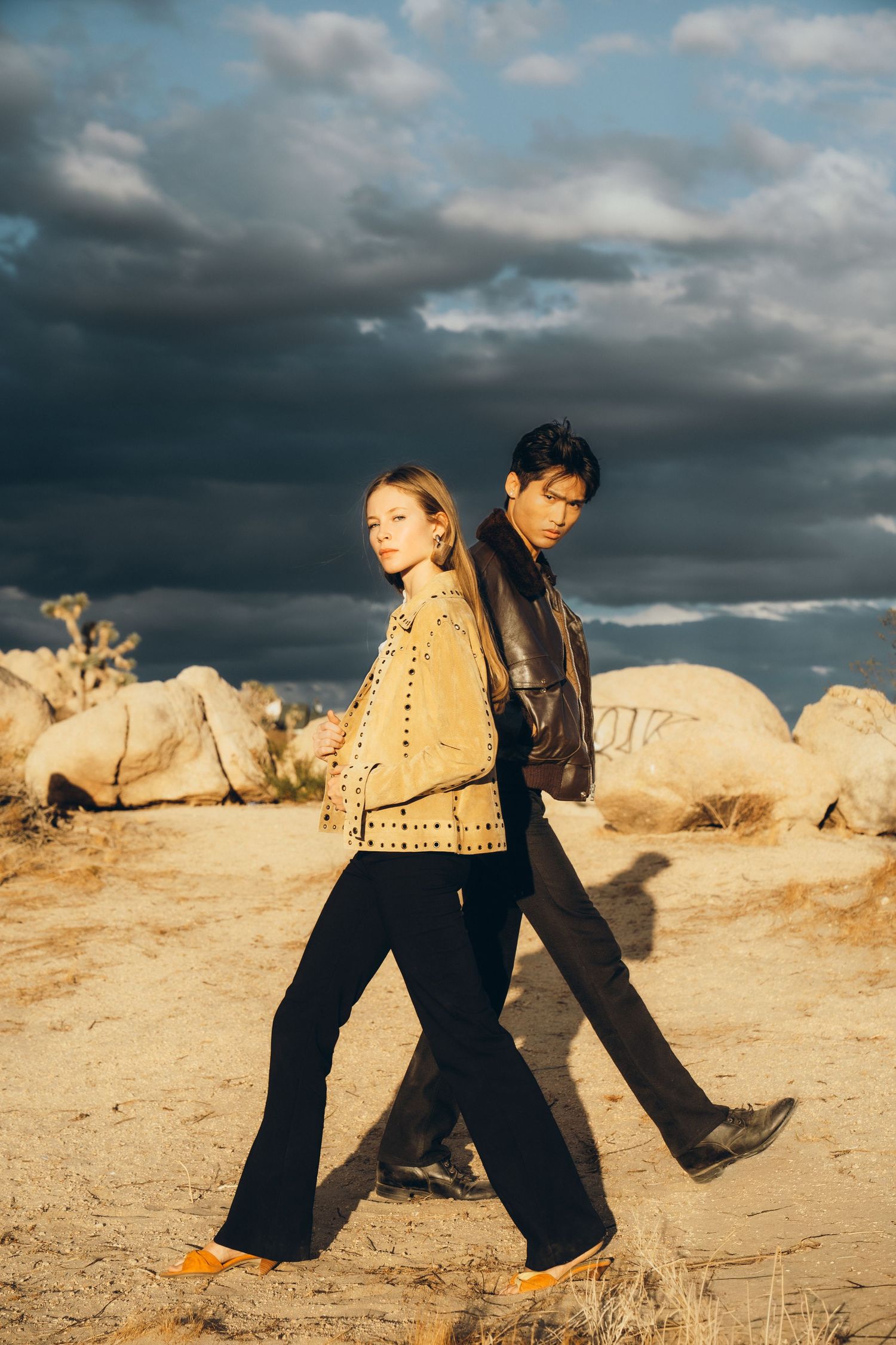 Two people in black outfits dance across a desert landscape under dramatic cloudy skies at sunset.