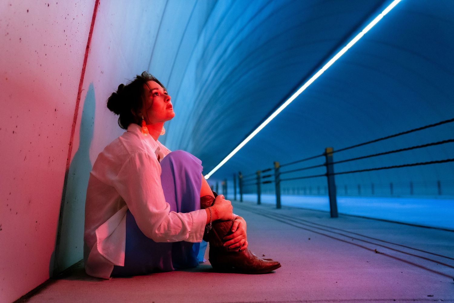 Person in pink jacket sits against wall illuminated by dramatic blue and pink lighting in tunnel-like space.