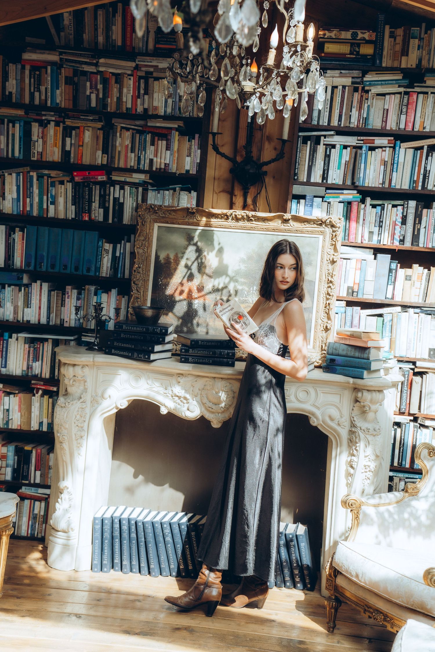 Woman in gray slip dress holding ornate gold mirror in a cozy library with floor-to-ceiling bookshelves and fireplace.