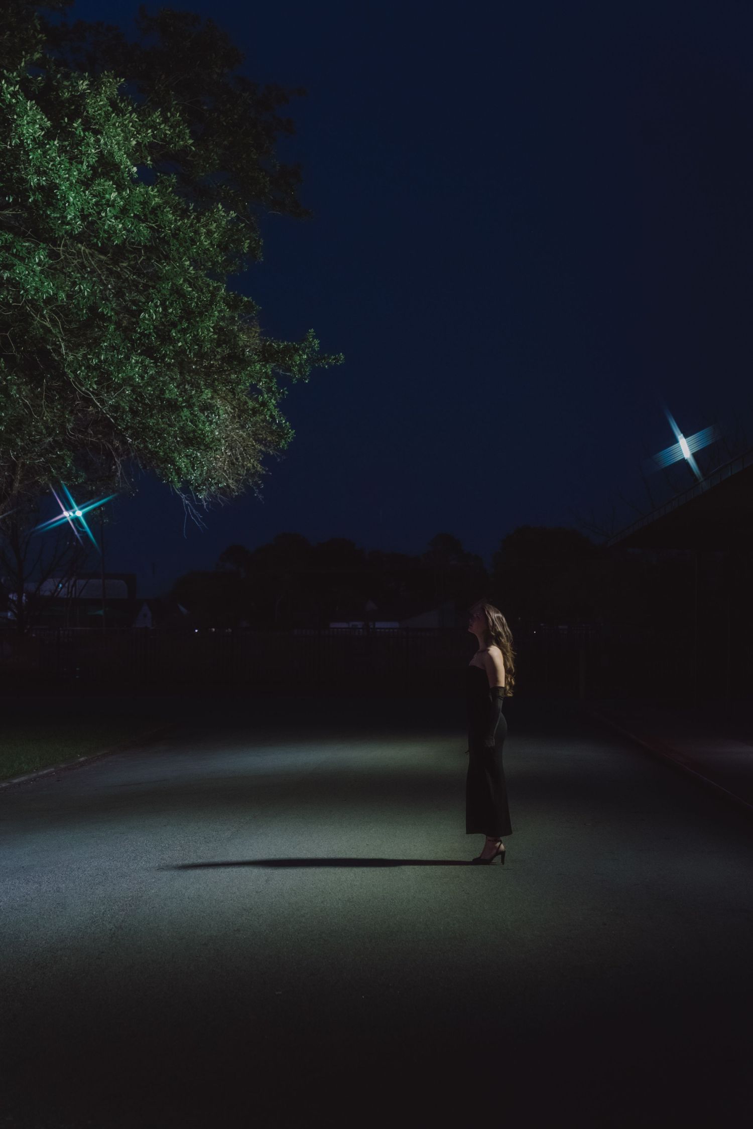 A figure illuminated by a flashlight stands alone in a dark outdoor setting under trees at night.