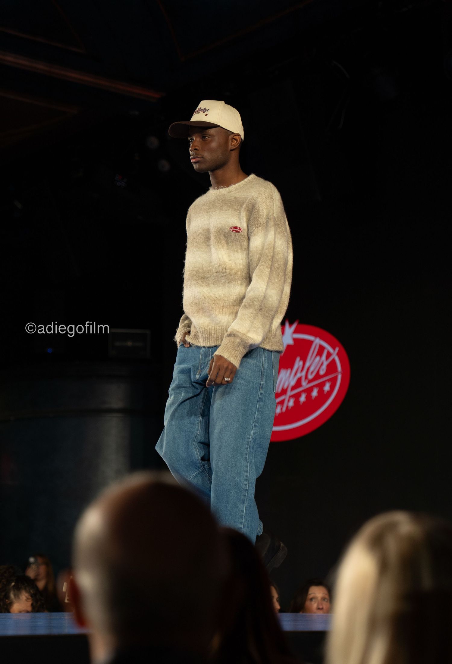 Fashion model wearing cream sweater and jeans walks runway with red neon sign in background during fashion show.