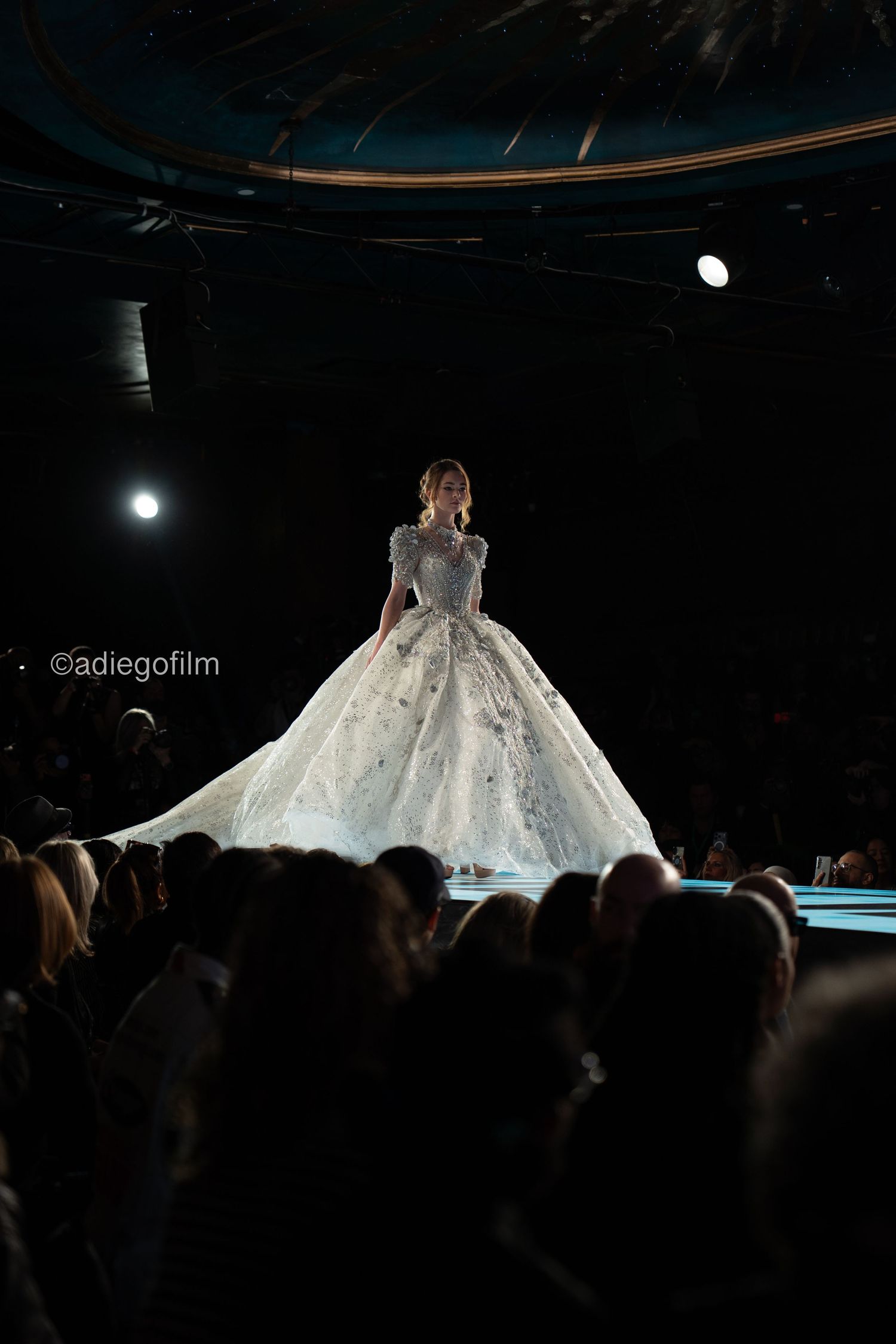 A stunning ball gown wedding dress sparkles on a darkened runway as audience members watch from the shadows.