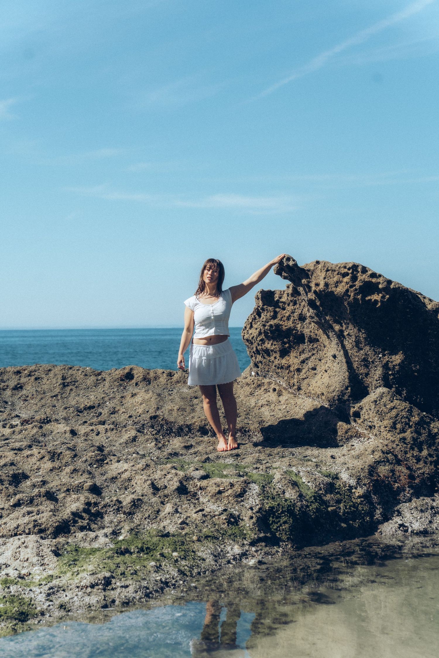 Woman in white outfit standing on rocky coastal shore with turquoise ocean and blue sky in background.