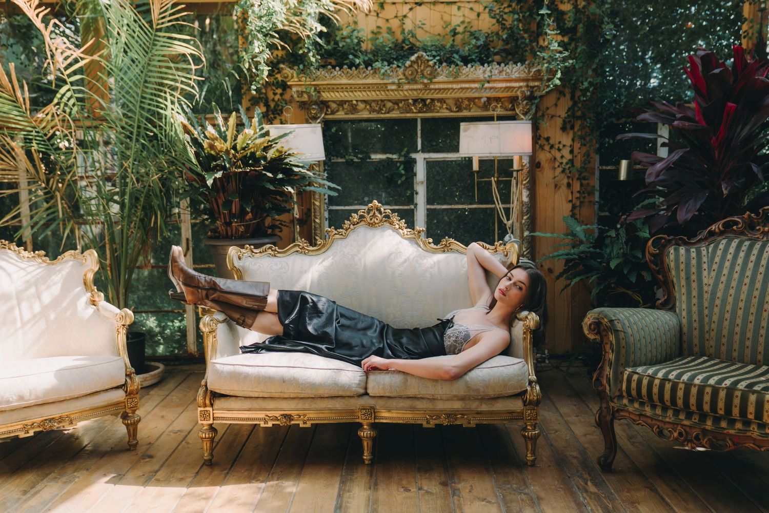 Person lounging on ornate gold-framed white sofa surrounded by lush tropical plants in an elegant vintage-style conservatory.