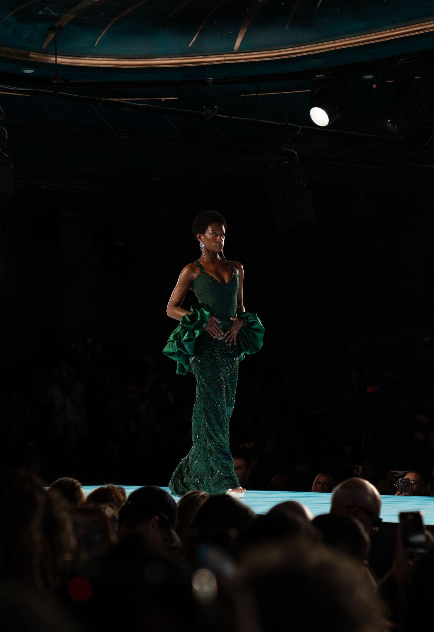 A performer in a green dress stands on stage silhouetted against dramatic lighting while an audience watches from below.