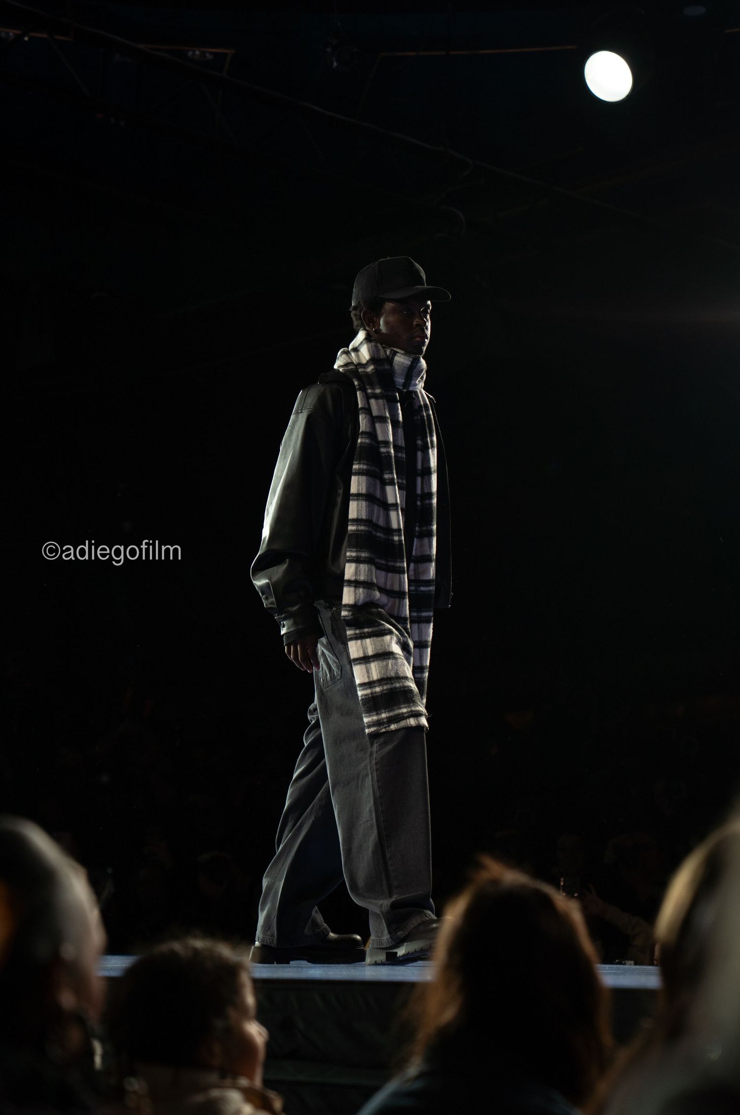 Fashion models walk the runway in dark lighting wearing plaid patterns and casual wear during a fashion show.