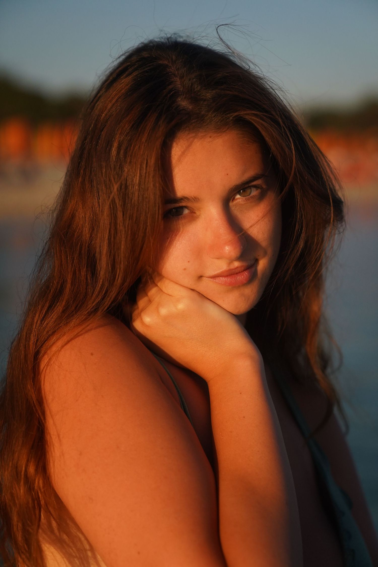 Portrait of long haired brunette in golden sunset light resting chin on hand while looking at camera near water.
