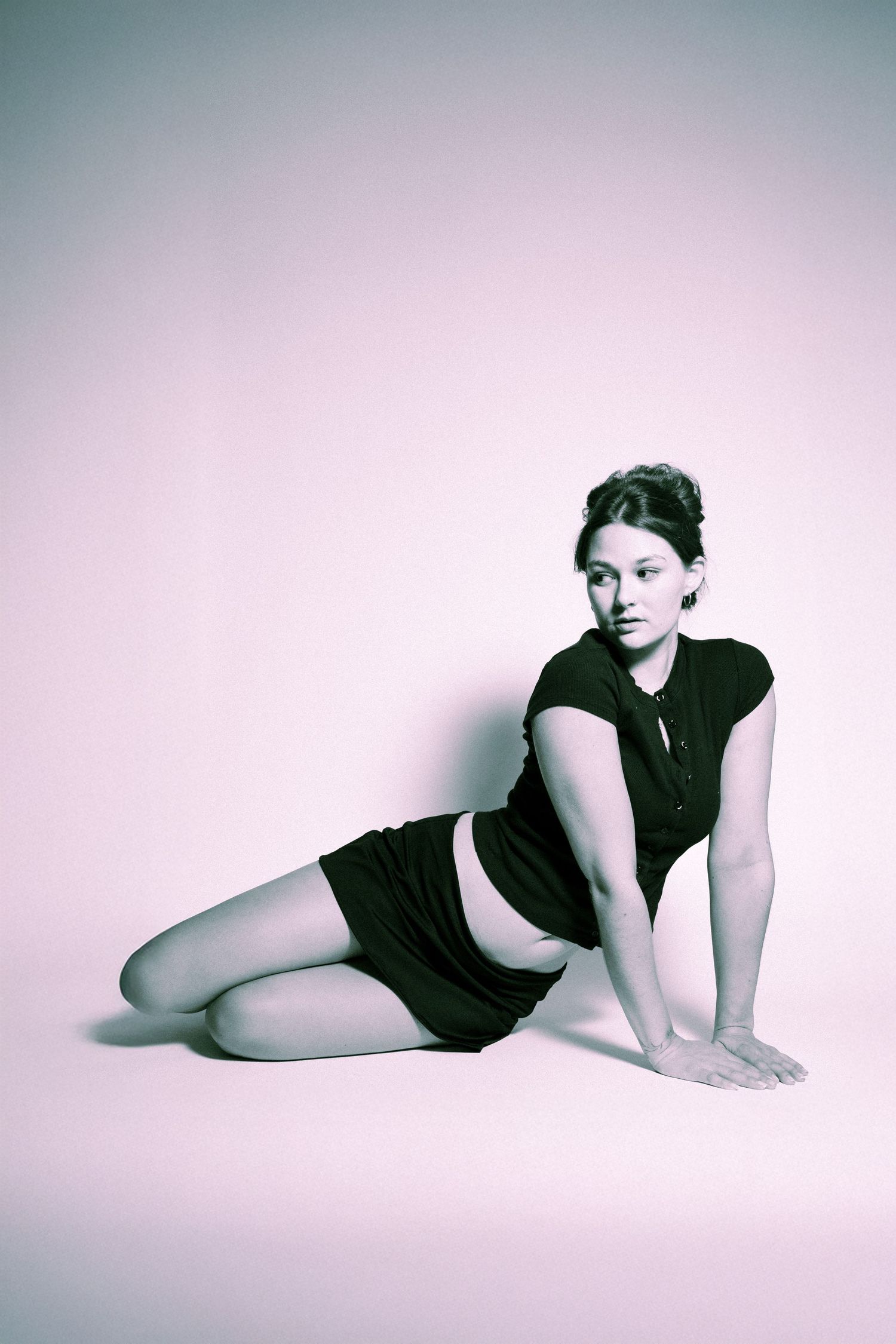 A model in a black dress poses in a reclining position against a pink gradient background in a stylized studio portrait.