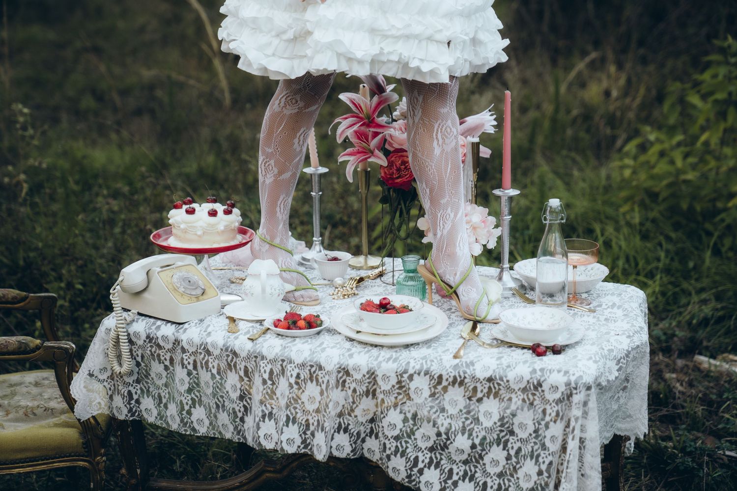 A rustic outdoor wedding dessert table with lace tablecloth, tiered cake, and floral decorations in a garden setting.