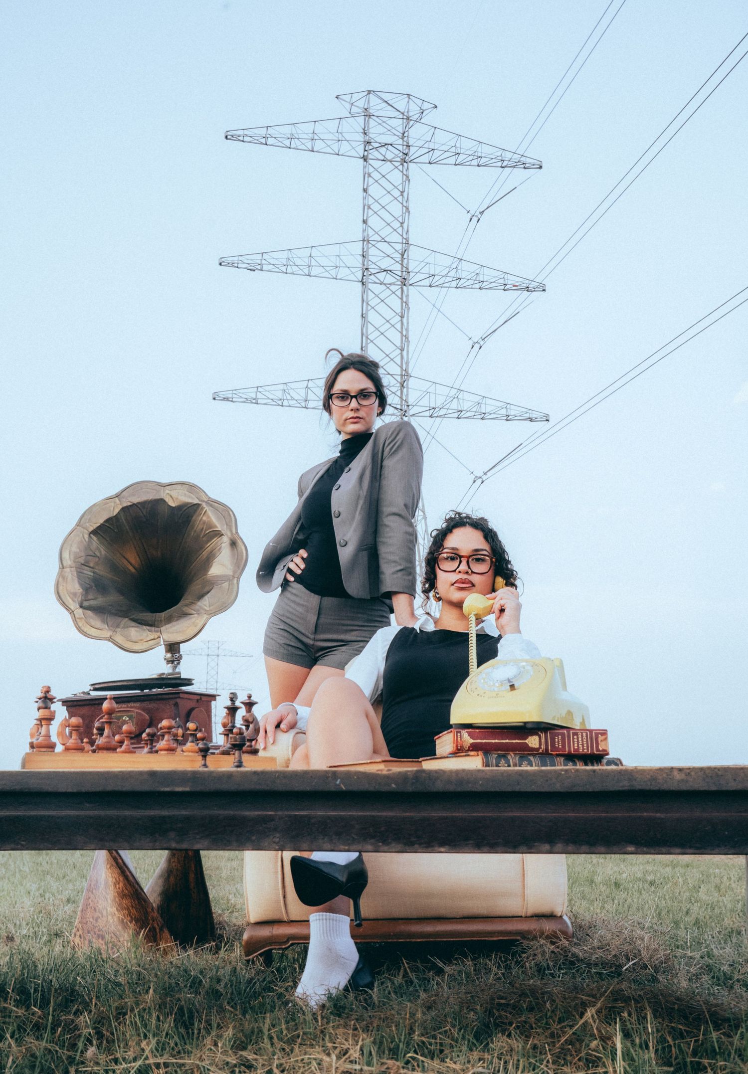 A vintage phonograph and chess set arranged on a wooden bench against a backdrop of power lines in a grassy field.