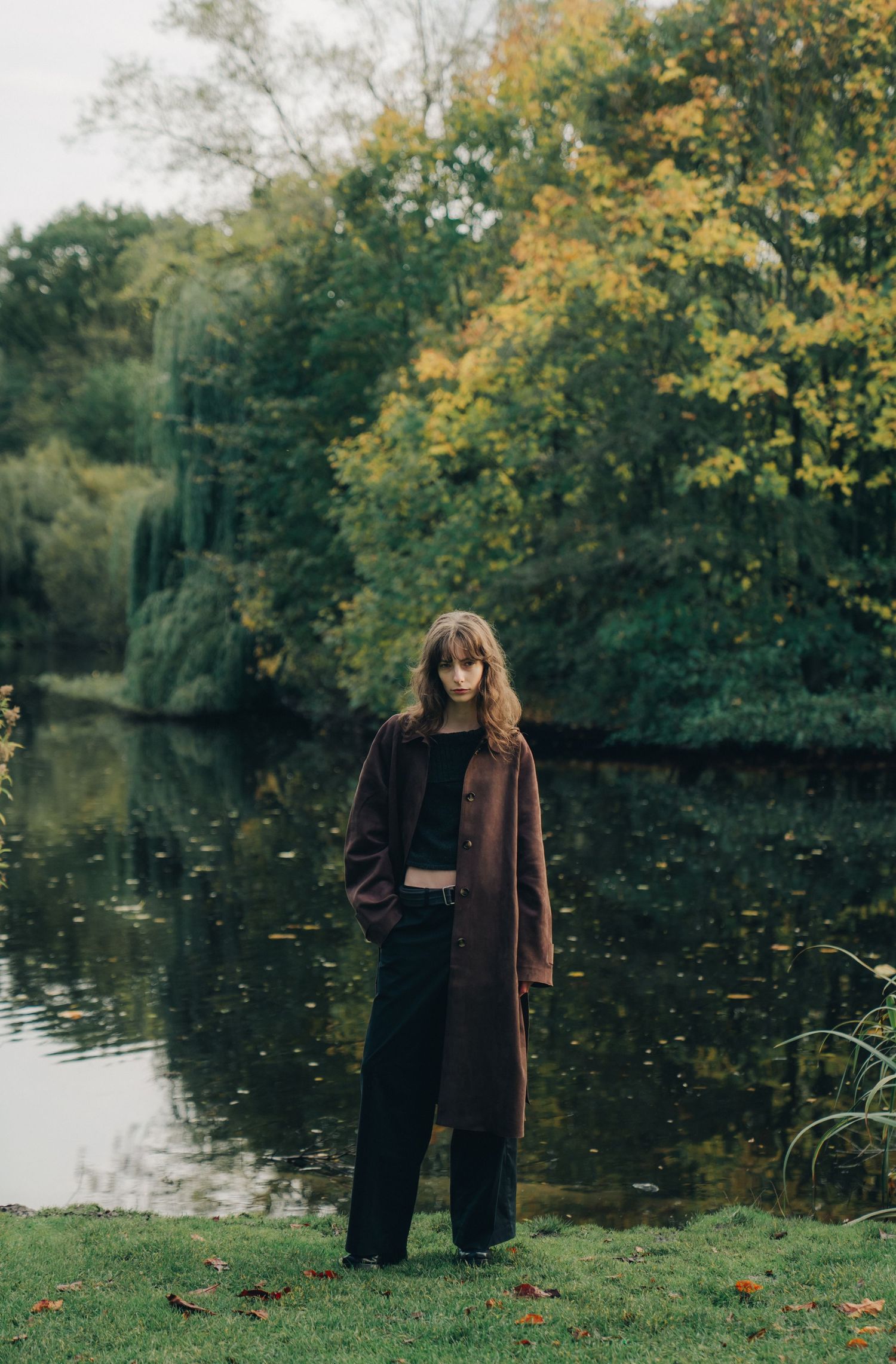 A figure in a brown coat and dark pants stands by a tranquil pond surrounded by autumn trees with yellow foliage.