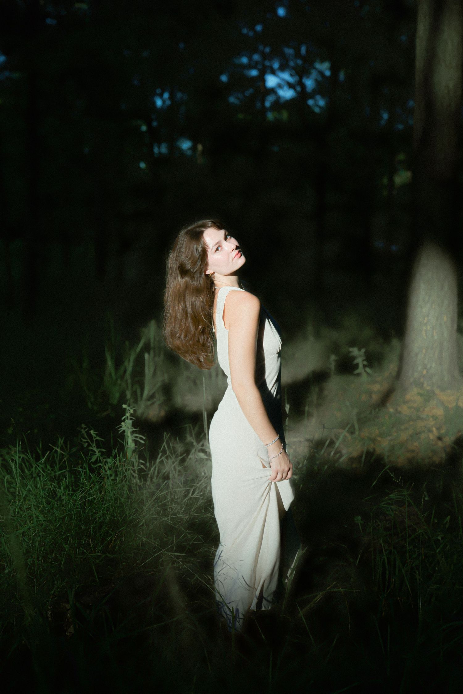 A woman in a white dress standing in a dark forest with soft lighting and dreamy atmosphere.