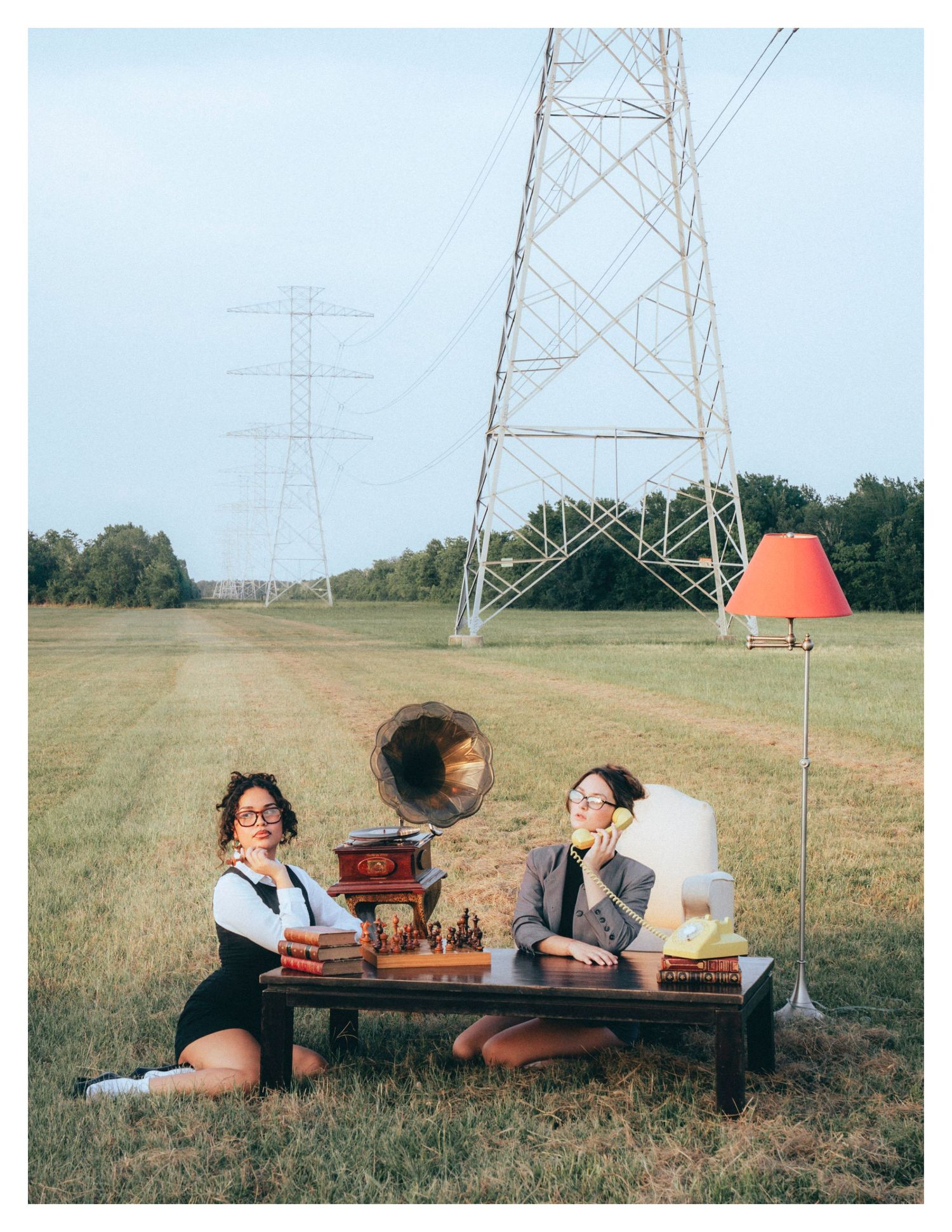 Two people sit at a low table with vintage decor under power lines in a grassy field during a retro photoshoot.